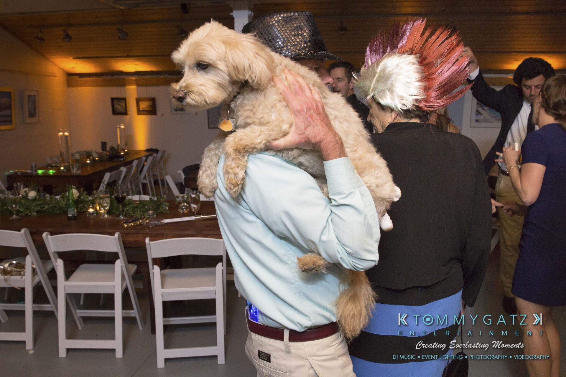 Amanda-Tyler-102117-RECEPTION-Low-Res-WM-202 - TOMMY GATZ ENTERTAINMENT A man in a party setting holds a dog on his shoulder. People are socializing in the background, and the room is decorated with tables and chairs.