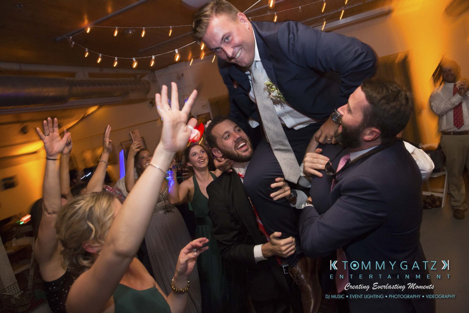 Amanda-Tyler-102117-RECEPTION-Low-Res-WM-213 - TOMMY GATZ ENTERTAINMENT Group of people celebrating indoors, with one person being lifted on others' shoulders. String lights are in the background.