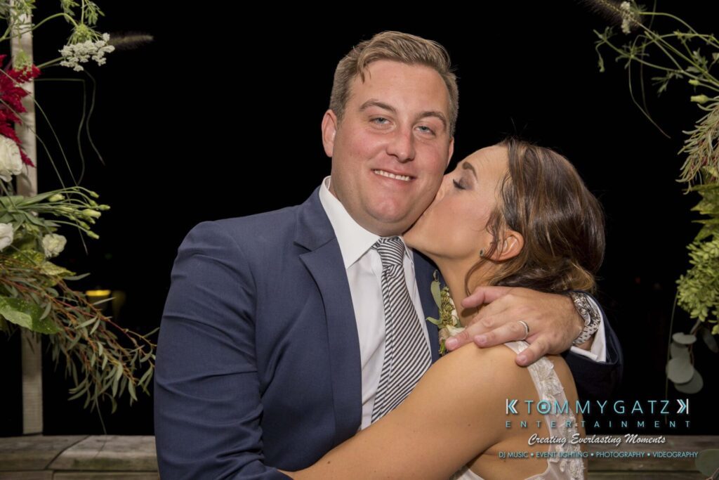 A person in a suit receives a kiss on the cheek from a woman in a white dress, set against a floral backdrop.
