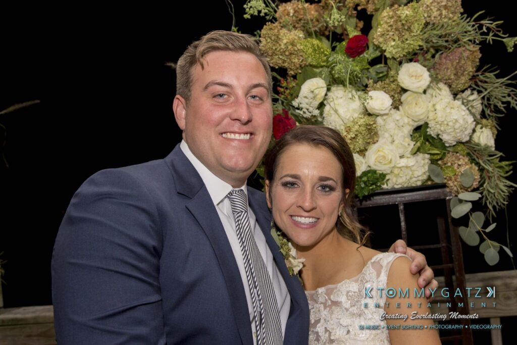 Amanda-Tyler-102117-RECEPTION-Low-Res-WM-225 - TOMMY GATZ ENTERTAINMENT A couple dressed in formal attire poses together, smiling in front of a floral arrangement.