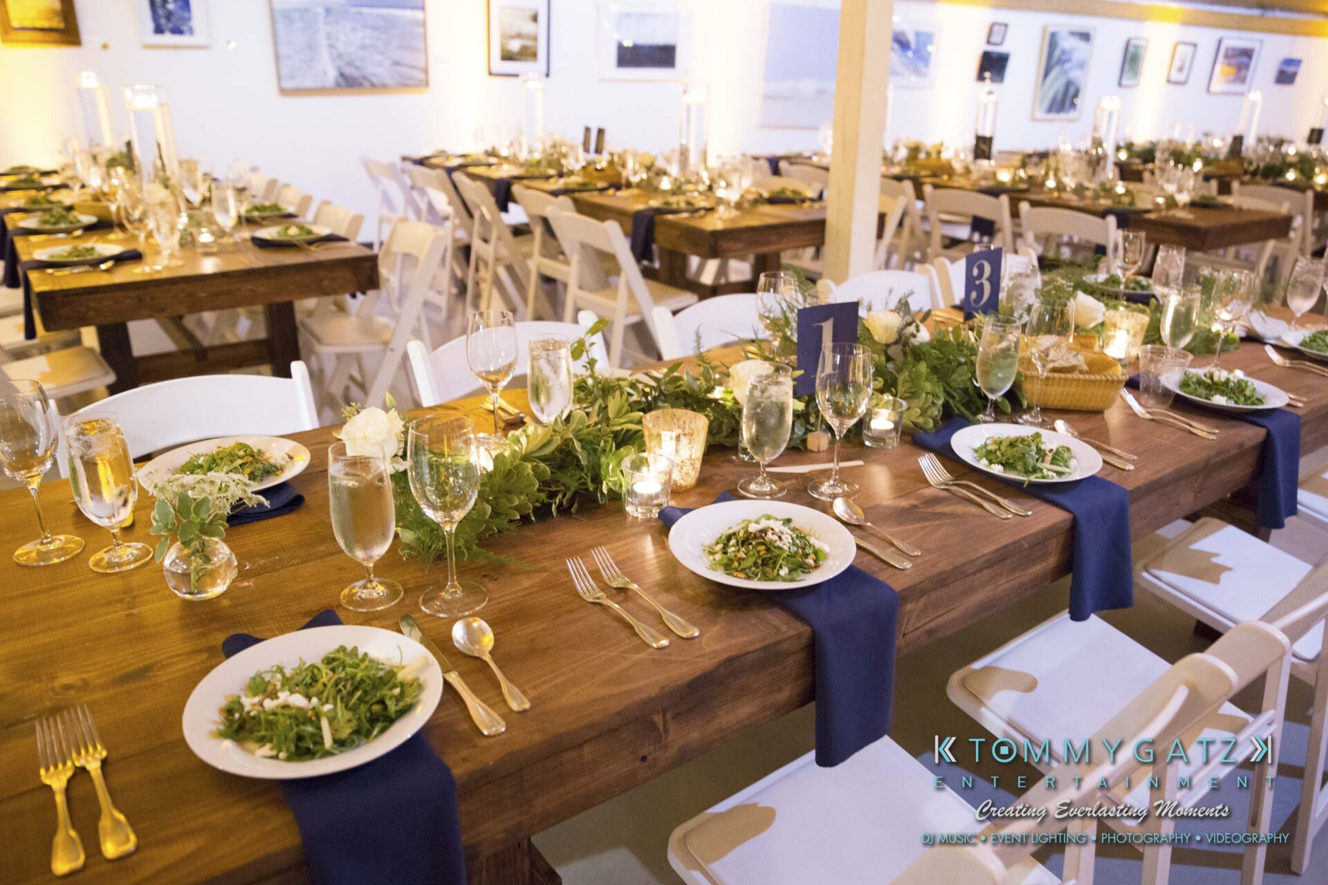 Amanda-Tyler-102117-RECEPTION-Low-Res-WM-25 - TOMMY GATZ ENTERTAINMENT A dining setup with long wooden tables, white chairs, and place settings featuring salads and drinks. Greenery and candles decorate the tables, creating an elegant atmosphere.