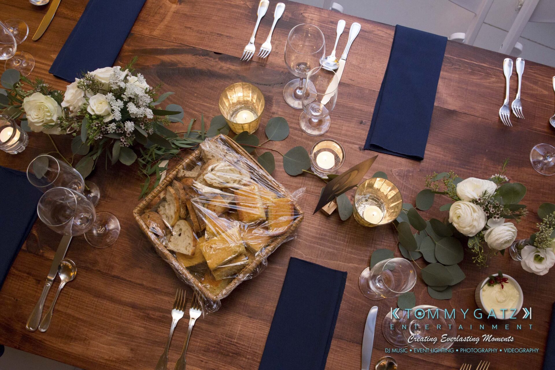 Amanda-Tyler-102117-RECEPTION-Low-Res-WM-3 - TOMMY GATZ ENTERTAINMENT A wooden table set for a meal with a basket of bread wrapped in plastic, white roses, candles, navy napkins, and glasses.
