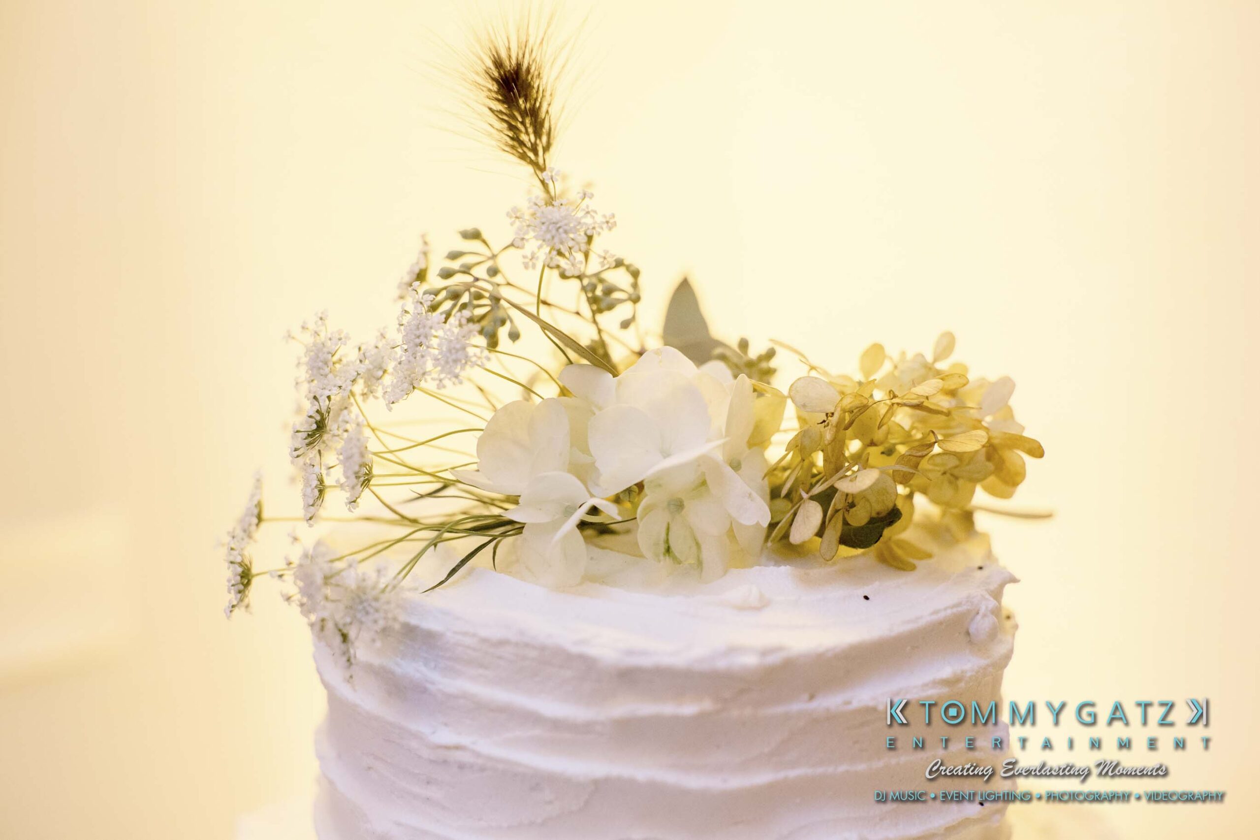 Amanda-Tyler-102117-RECEPTION-Low-Res-WM-36 - TOMMY GATZ ENTERTAINMENT A white frosted cake topped with white and pale yellow flowers, and a single stalk of wheat.