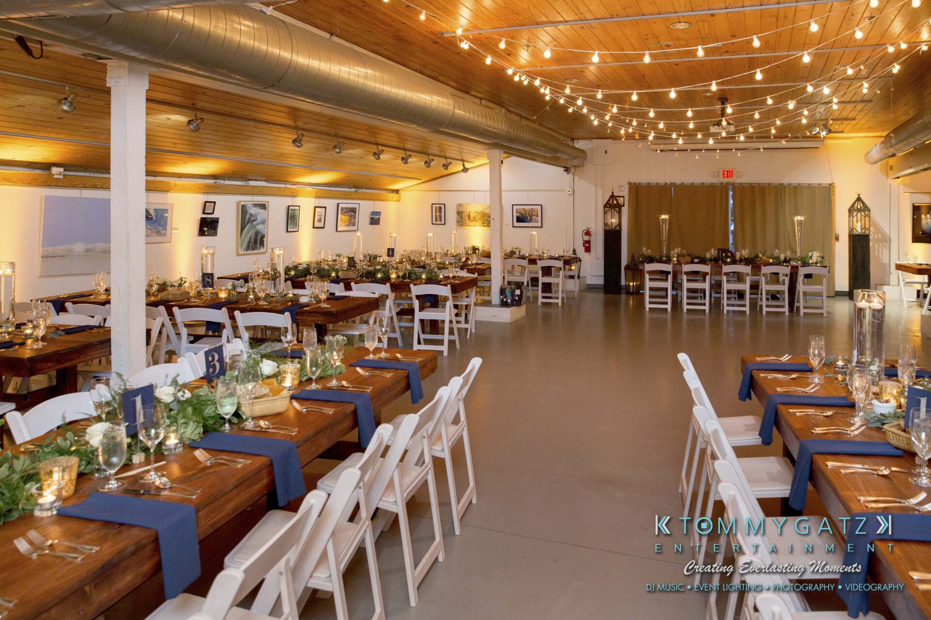 Amanda-Tyler-102117-RECEPTION-Low-Res-WM-4 - TOMMY GATZ ENTERTAINMENT A decorated banquet hall with wooden tables, white chairs, and table centerpieces. The room has artwork on the walls and a wooden ceiling.