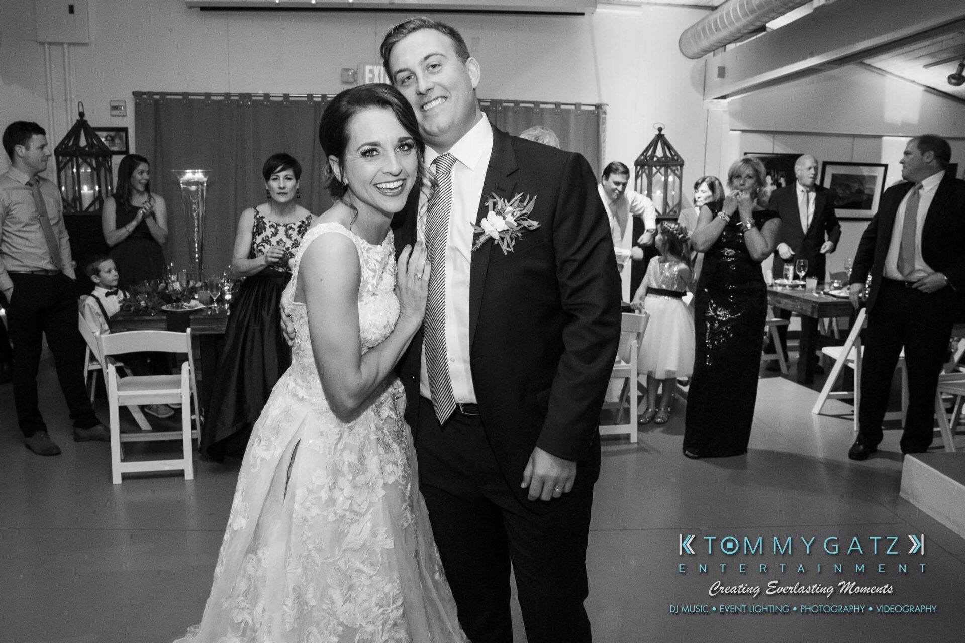 Amanda-Tyler-102117-RECEPTION-Low-Res-WM-43 - TOMMY GATZ ENTERTAINMENT A bride and groom smile for a photo at a wedding reception, surrounded by guests seated and standing in the background.