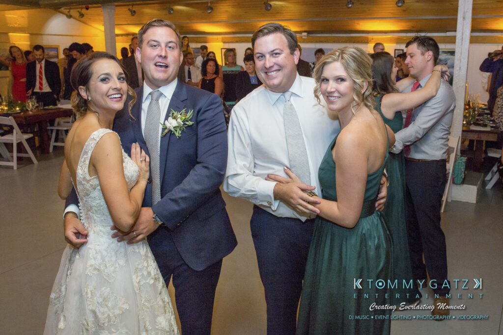 Amanda-Tyler-102117-RECEPTION-Low-Res-WM-47 - TOMMY GATZ ENTERTAINMENT Two couples pose and smile for a photo at an indoor event. Guests and tables are visible in the background.