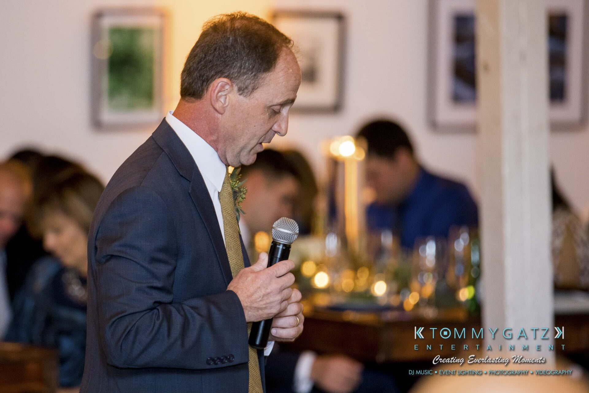 Amanda-Tyler-102117-RECEPTION-Low-Res-WM-48 - TOMMY GATZ ENTERTAINMENT A man in a suit speaks into a microphone at an indoor event with seated guests in the background.