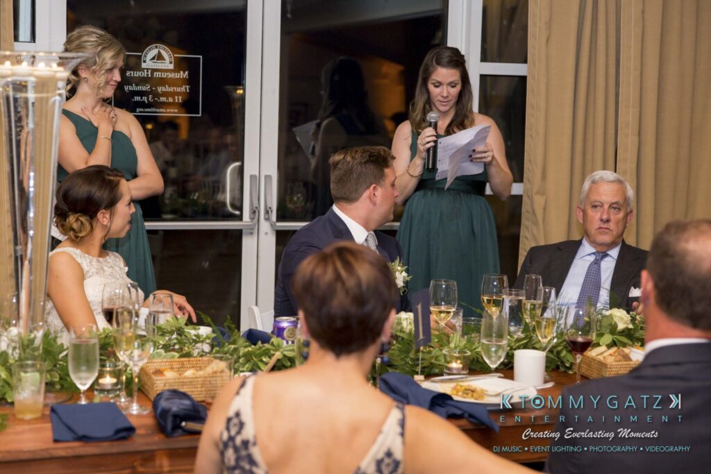 Amanda-Tyler-102117-RECEPTION-Low-Res-WM-54 - TOMMY GATZ ENTERTAINMENT A woman in a green dress gives a speech at a dinner event. Five people sit around a table set with drinks and decor.
