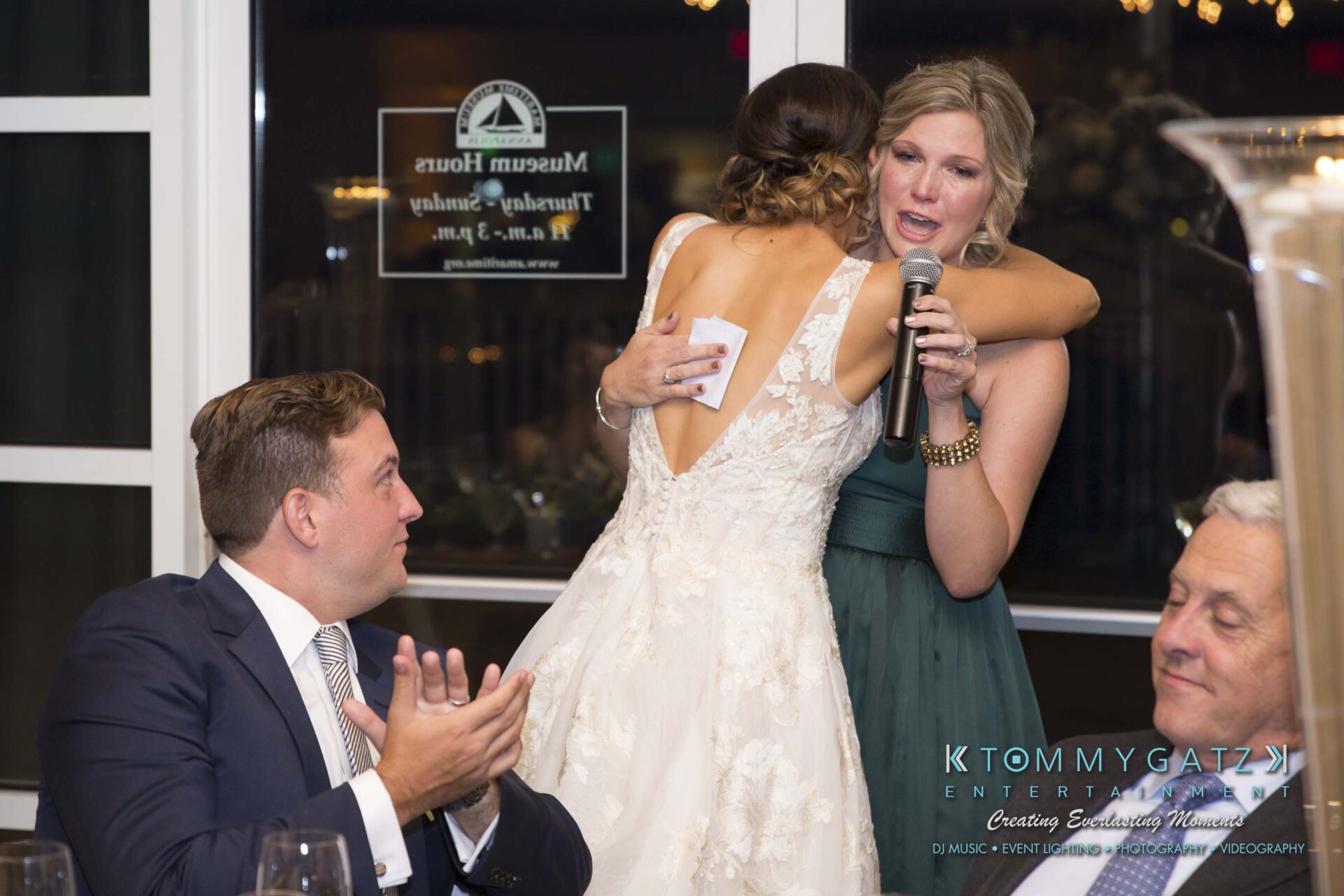 Amanda-Tyler-102117-RECEPTION-Low-Res-WM-60 - TOMMY GATZ ENTERTAINMENT A bride hugs a woman holding a microphone at a wedding reception. A man in a suit claps in the foreground. Guests are seated at tables.