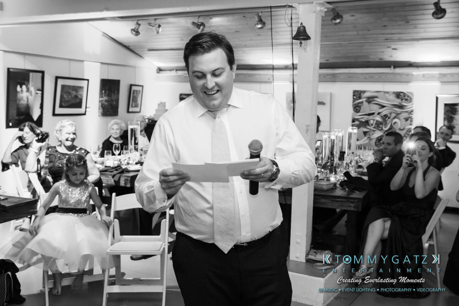 Amanda-Tyler-102117-RECEPTION-Low-Res-WM-61 - TOMMY GATZ ENTERTAINMENT A man in a tie speaks into a microphone while holding a paper, with guests seated around tables in a decorated room.
