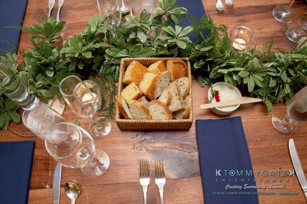 Basket of assorted bread on a wooden table with blue napkins, wine glasses, candles, and greenery decoration.