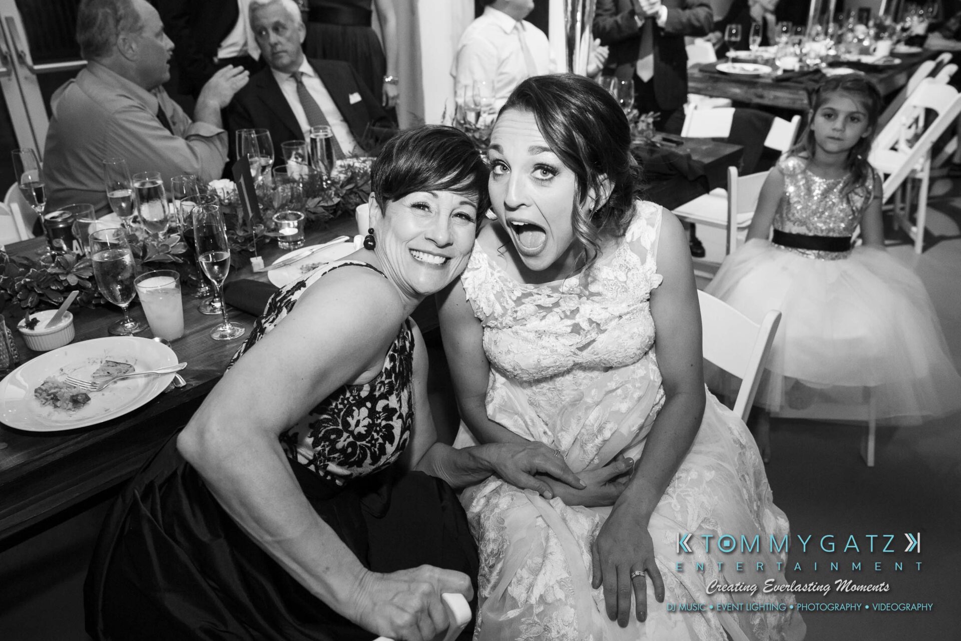 Amanda-Tyler-102117-RECEPTION-Low-Res-WM-78 - TOMMY GATZ ENTERTAINMENT Two women smiling, seated at a table set for a formal event. One is wearing a textured dress, the other in a white gown. A child and other attendees are visible in the background.