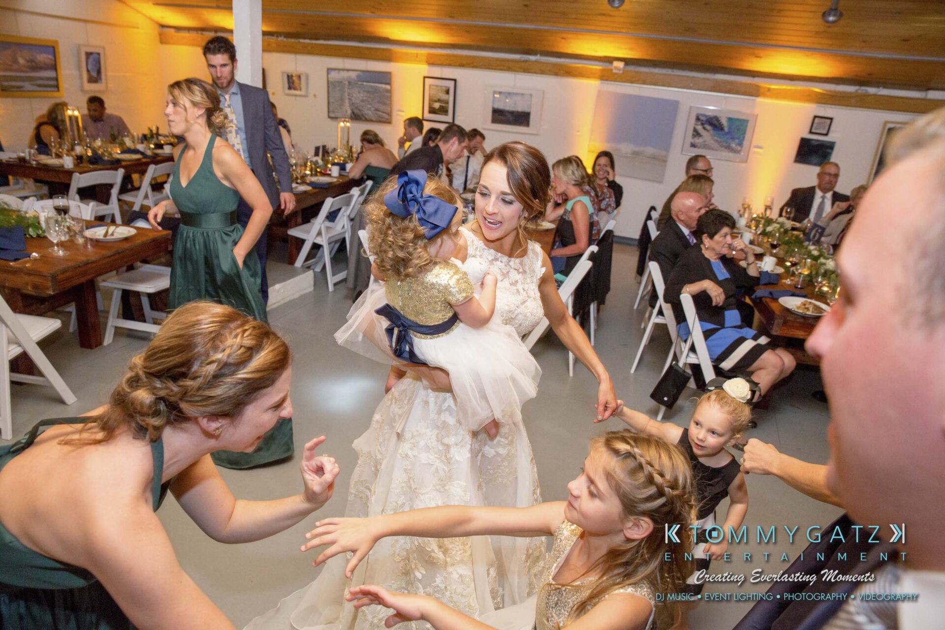 Amanda-Tyler-102117-RECEPTION-Low-Res-WM-89 - TOMMY GATZ ENTERTAINMENT A wedding reception with guests dancing. A bride in a white dress holds a child. Tables are set with food and drinks in the background.