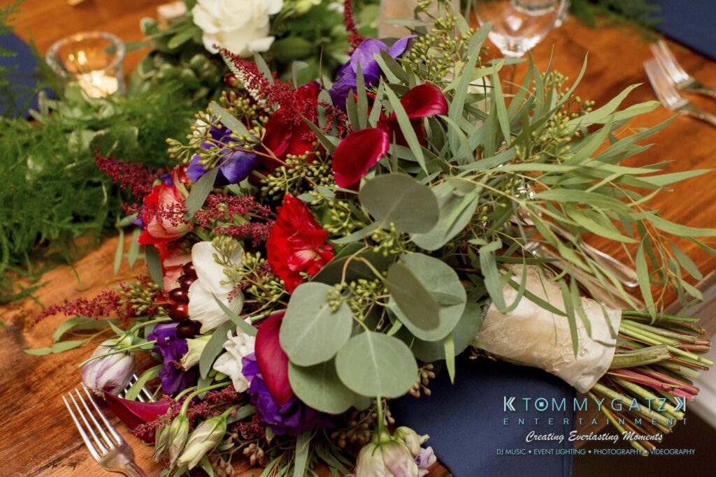 Amanda-Tyler-102117-RECEPTION-Low-Res-WM-9 - TOMMY GATZ ENTERTAINMENT A vibrant floral arrangement with red, purple, and white flowers, green eucalyptus leaves, and ferns on a wooden table. The logo "K Tommy Gatz Entertainment" is visible at the bottom right.
