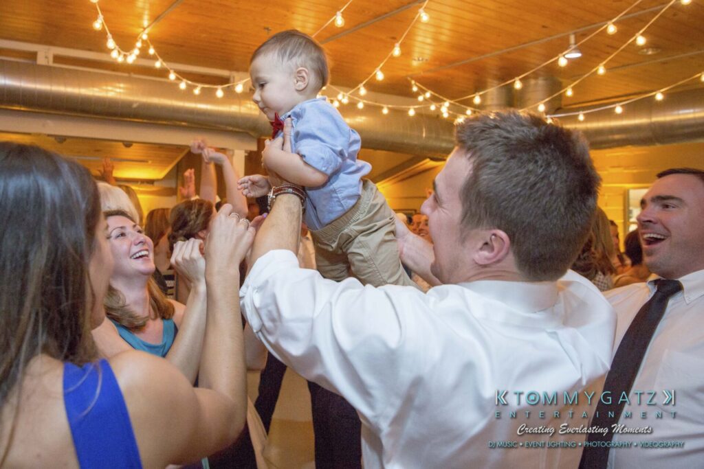 A group of people celebrating indoors, with a child being lifted into the air. String lights are hanging from the ceiling.