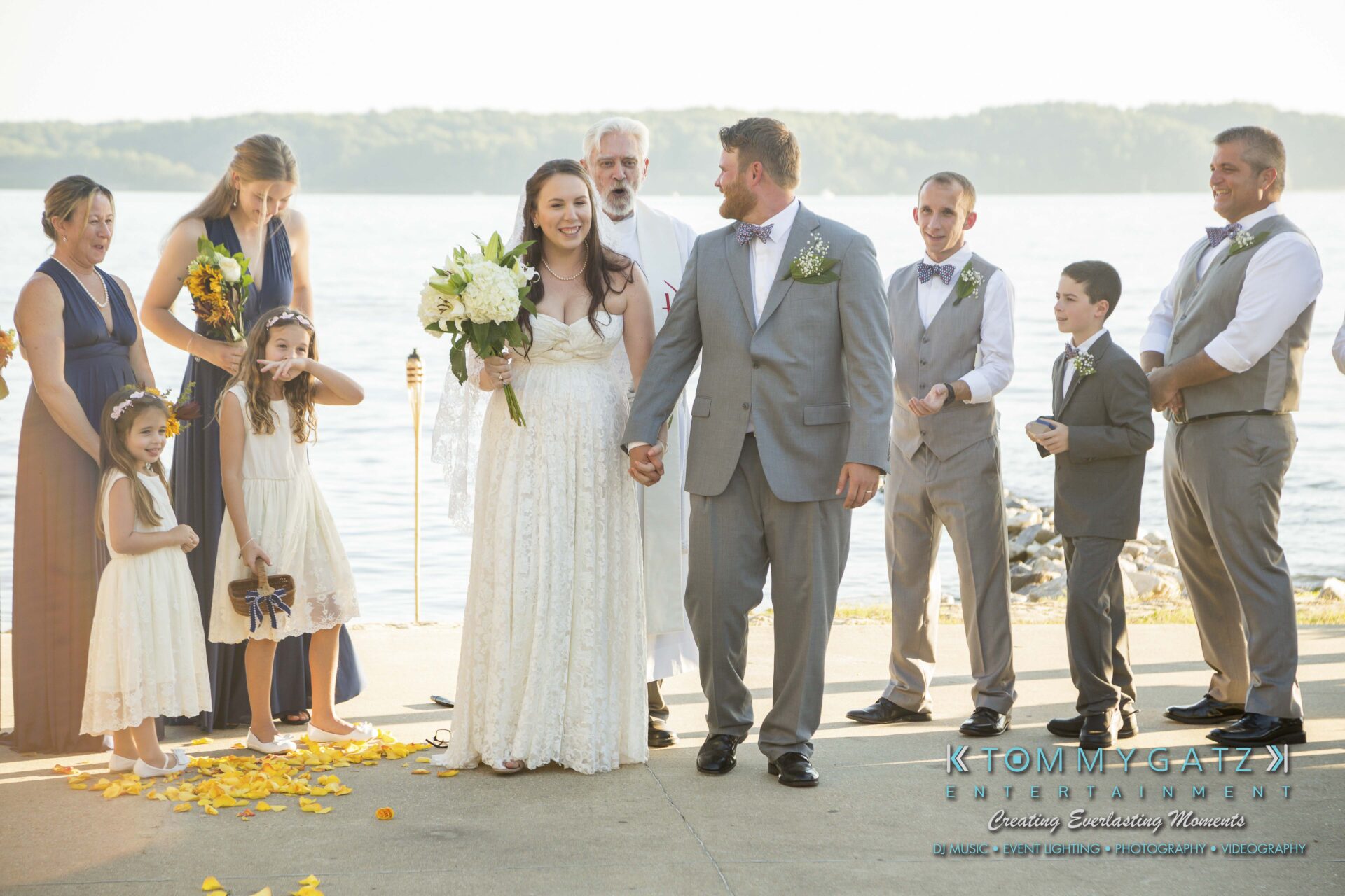 Ann-Marie-Alex-92317-CEREMONY-Low-Res-WM-102 - TOMMY GATZ ENTERTAINMENT A bride and groom stand outdoors near water, surrounded by their wedding party and family, including adults and children, dressed in formal attire.