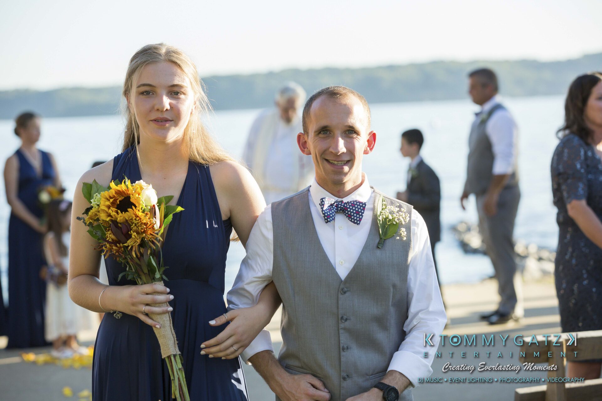 Ann-Marie-Alex-92317-CEREMONY-Low-Res-WM-106 - TOMMY GATZ ENTERTAINMENT A woman in a blue dress holds sunflowers, standing with a man in a gray vest and bow tie by a waterfront. Others are in the background.