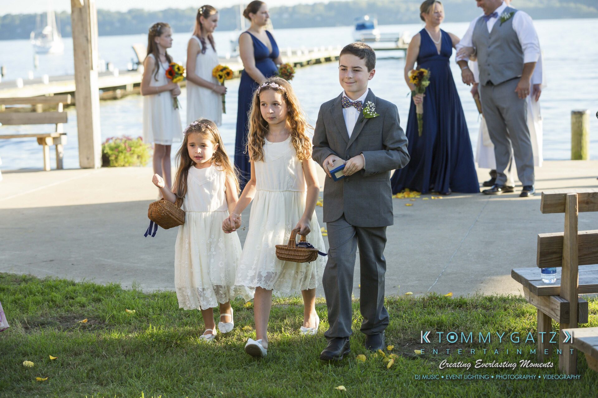 Ann-Marie-Alex-92317-CEREMONY-Low-Res-WM-107 - TOMMY GATZ ENTERTAINMENT Three children dressed in formal attire walk on a grassy area near a waterfront. The two girls wear white dresses and hold baskets, while the boy wears a gray suit. Adults in formal wear are in the background.