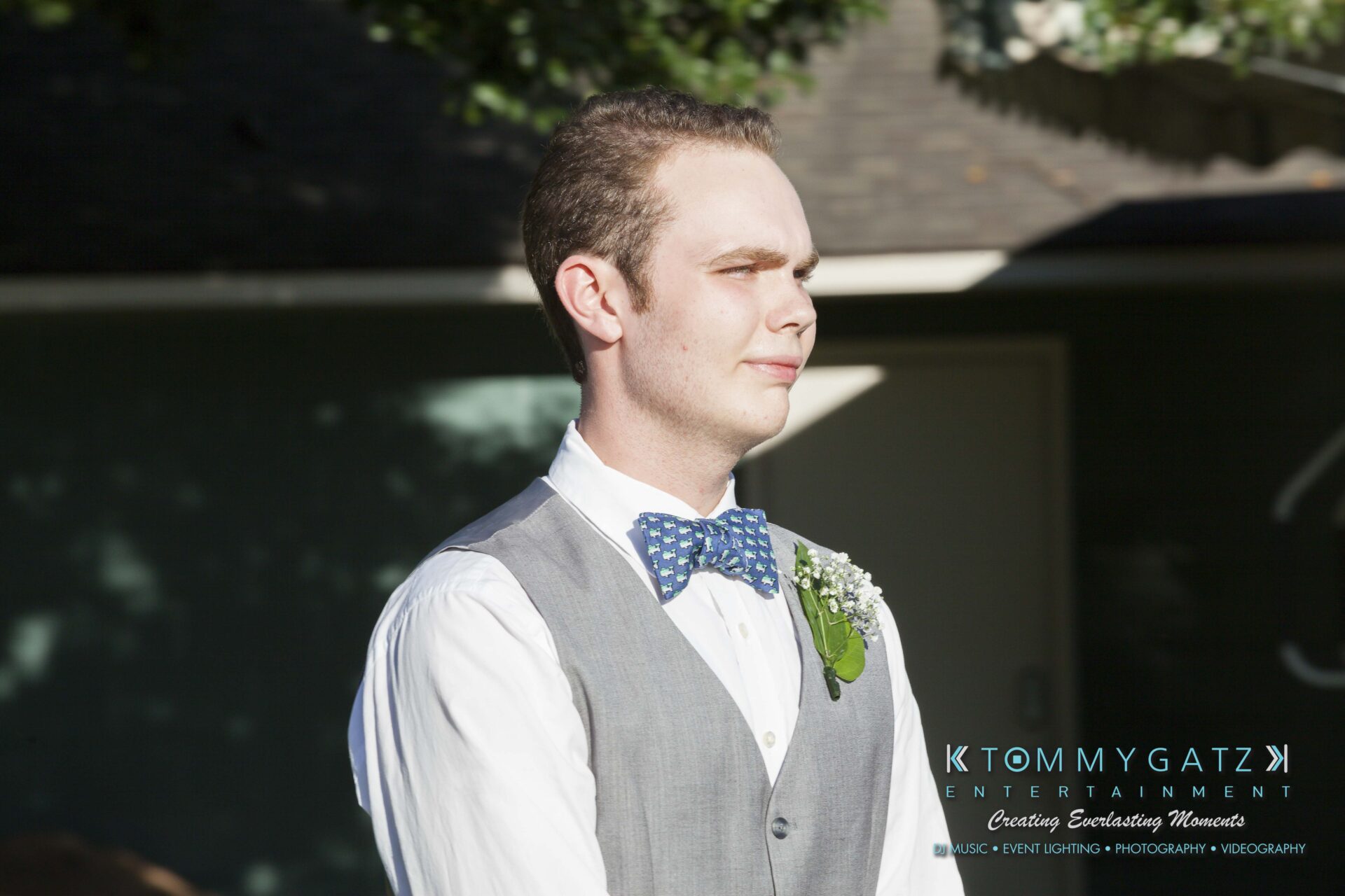 Ann-Marie-Alex-92317-CEREMONY-Low-Res-WM-12 - TOMMY GATZ ENTERTAINMENT A person in a gray vest and blue bow tie stands outdoors in sunlight, with a boutonniere on their left lapel.