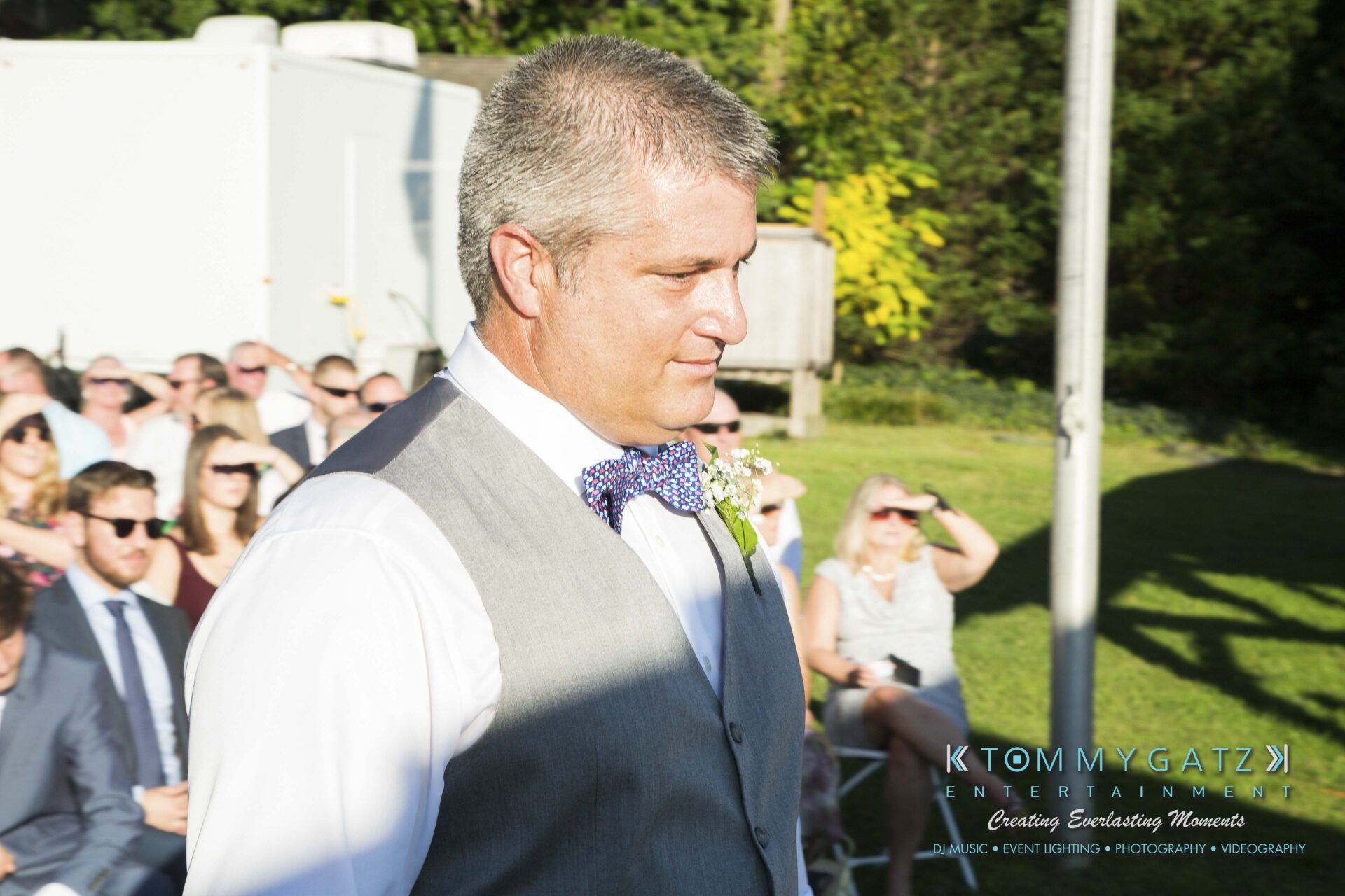Ann-Marie-Alex-92317-CEREMONY-Low-Res-WM-15 - TOMMY GATZ ENTERTAINMENT A man in a vest and tie walks outdoors during an event, with seated attendees in the background.