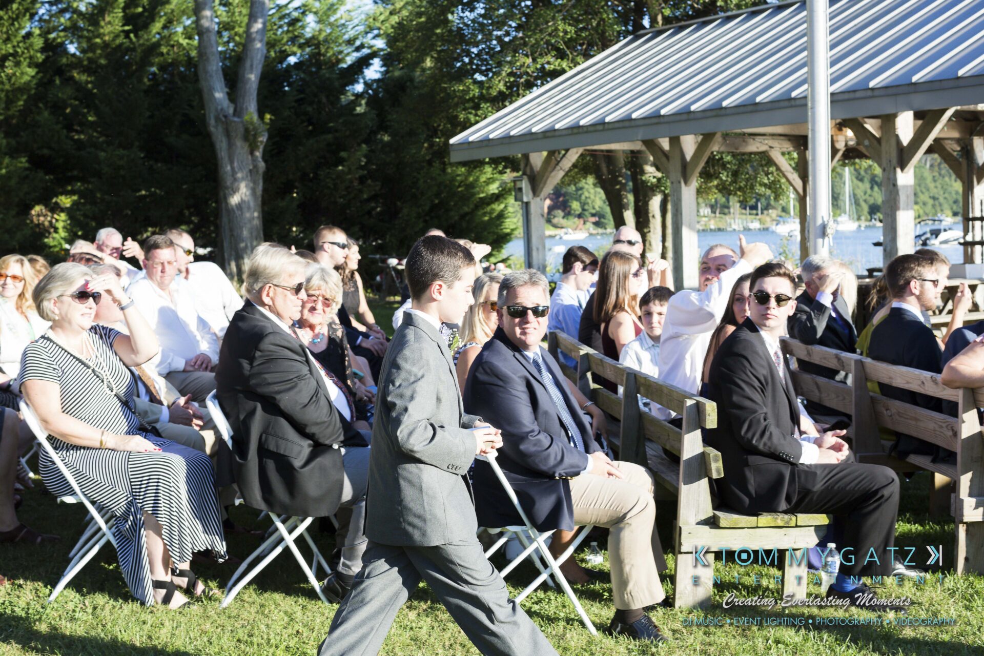 Ann-Marie-Alex-92317-CEREMONY-Low-Res-WM-16 - TOMMY GATZ ENTERTAINMENT A young boy in a suit walks past seated guests during an outdoor event on a sunny day.