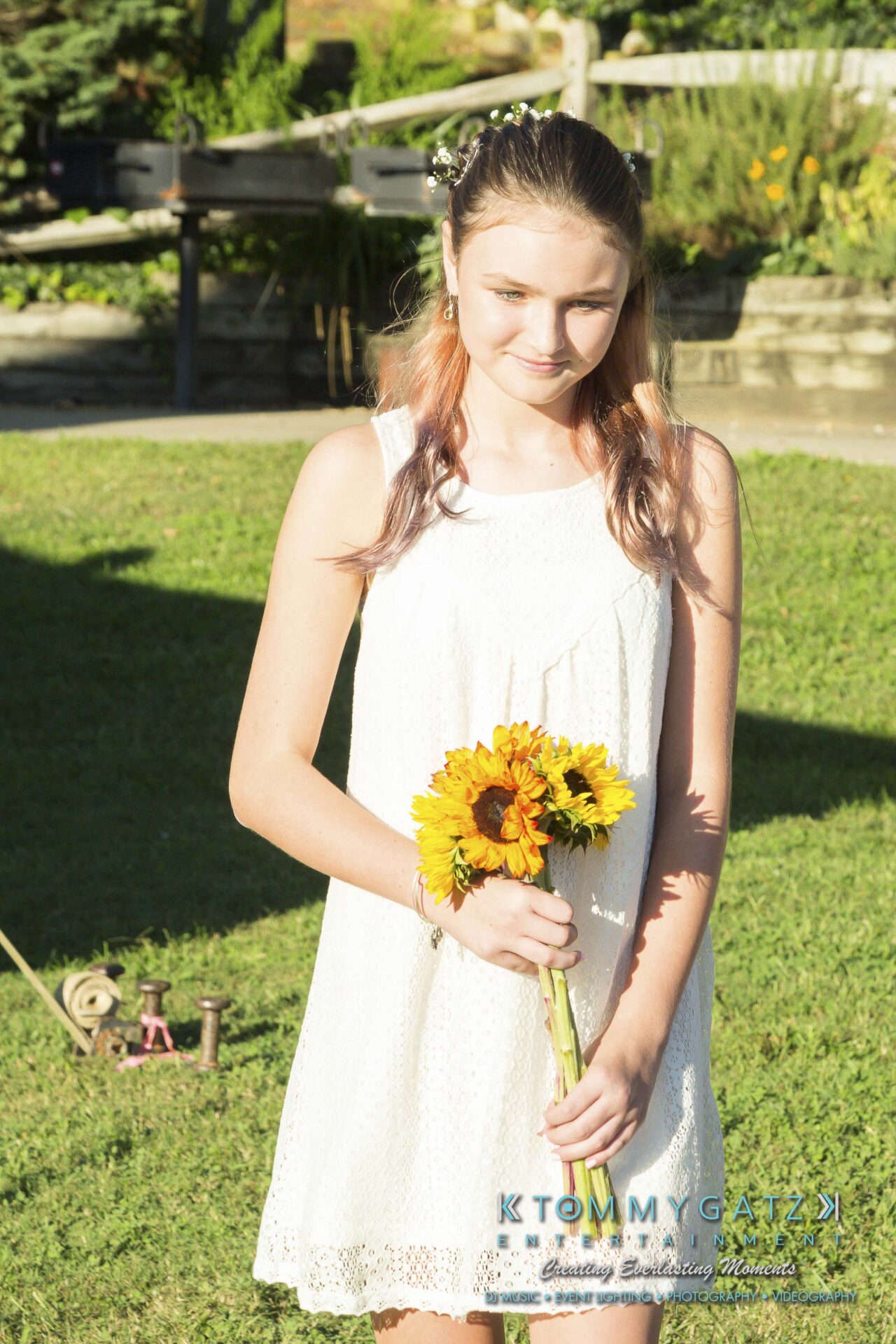 Ann-Marie-Alex-92317-CEREMONY-Low-Res-WM-19 - TOMMY GATZ ENTERTAINMENT Young woman in a white dress holding a bouquet of sunflowers, standing on grassy lawn with garden background.