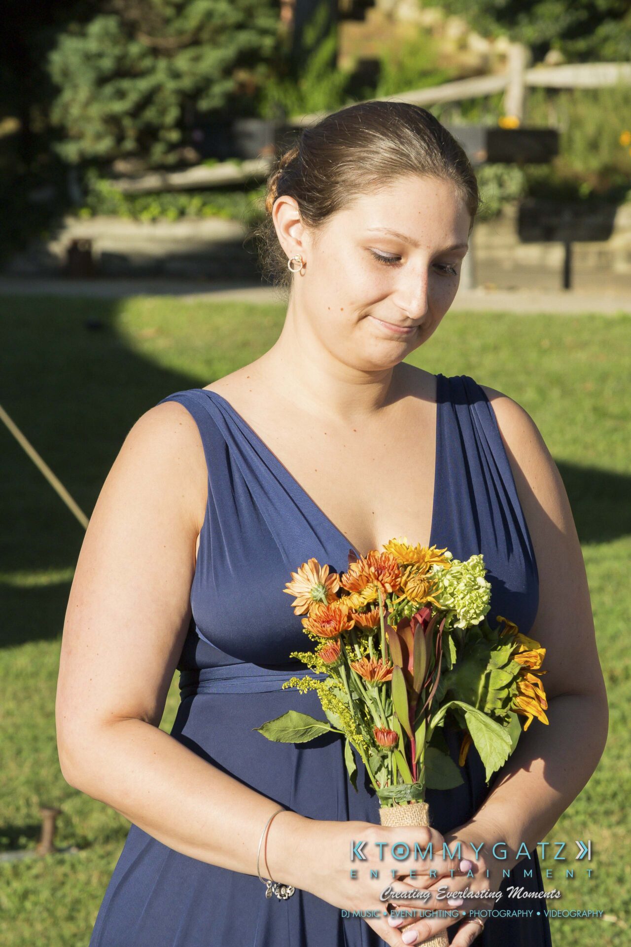 Ann-Marie-Alex-92317-CEREMONY-Low-Res-WM-21 - TOMMY GATZ ENTERTAINMENT A woman in a blue dress holds a bouquet of flowers while standing outdoors on grass.