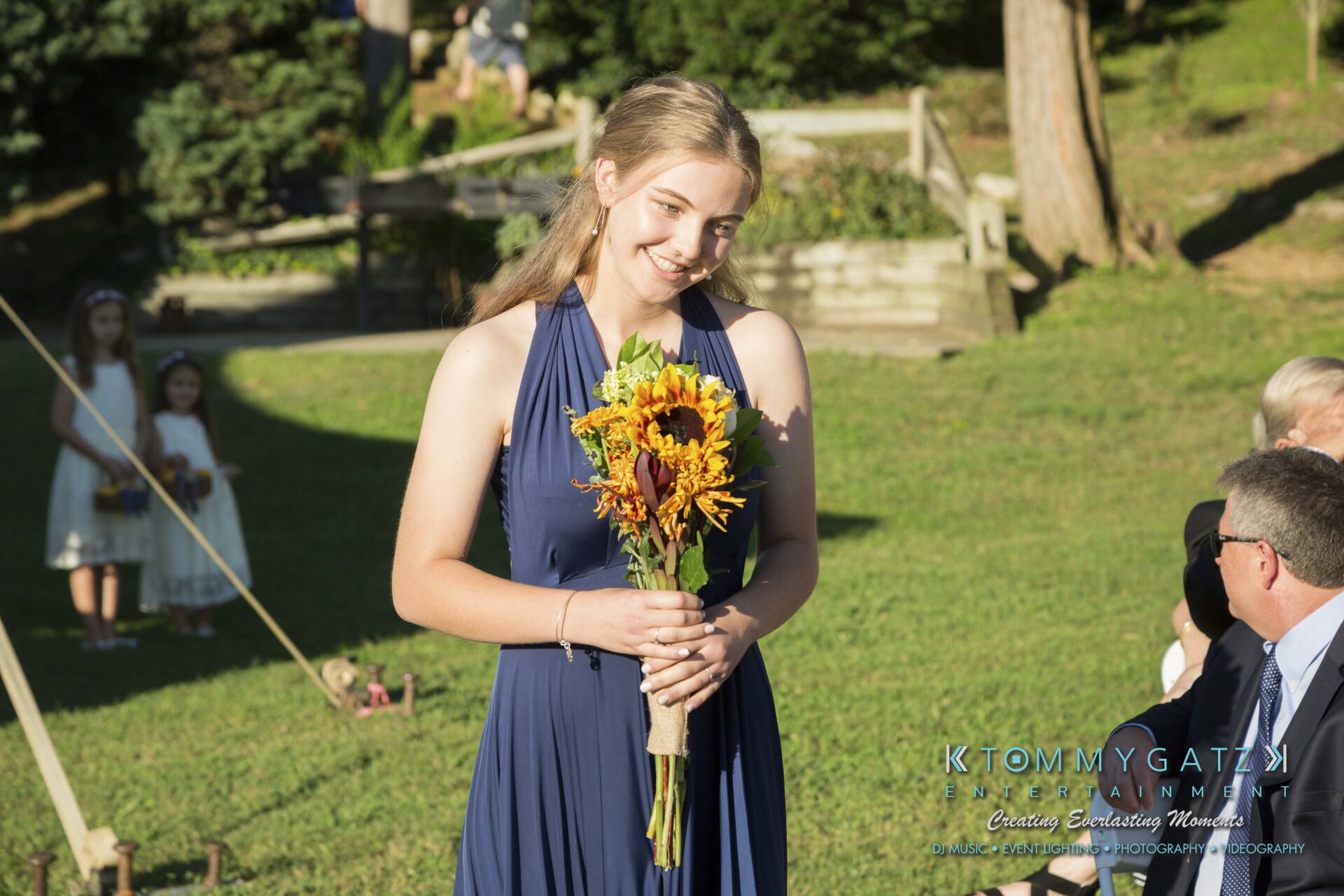 Ann-Marie-Alex-92317-CEREMONY-Low-Res-WM-25 - TOMMY GATZ ENTERTAINMENT A woman in a blue dress holds a bouquet of flowers while walking outdoors during a sunny event.