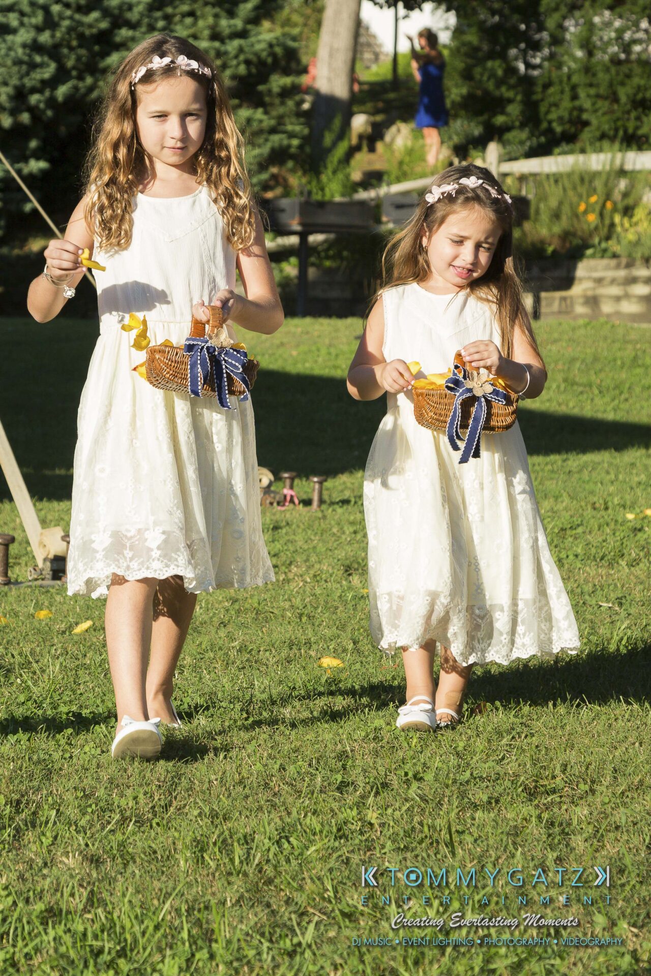 Ann-Marie-Alex-92317-CEREMONY-Low-Res-WM-28 - TOMMY GATZ ENTERTAINMENT Two young girls in white dresses walk on grass, holding baskets decorated with bows. They appear to be participating in a wedding or similar event outdoors.