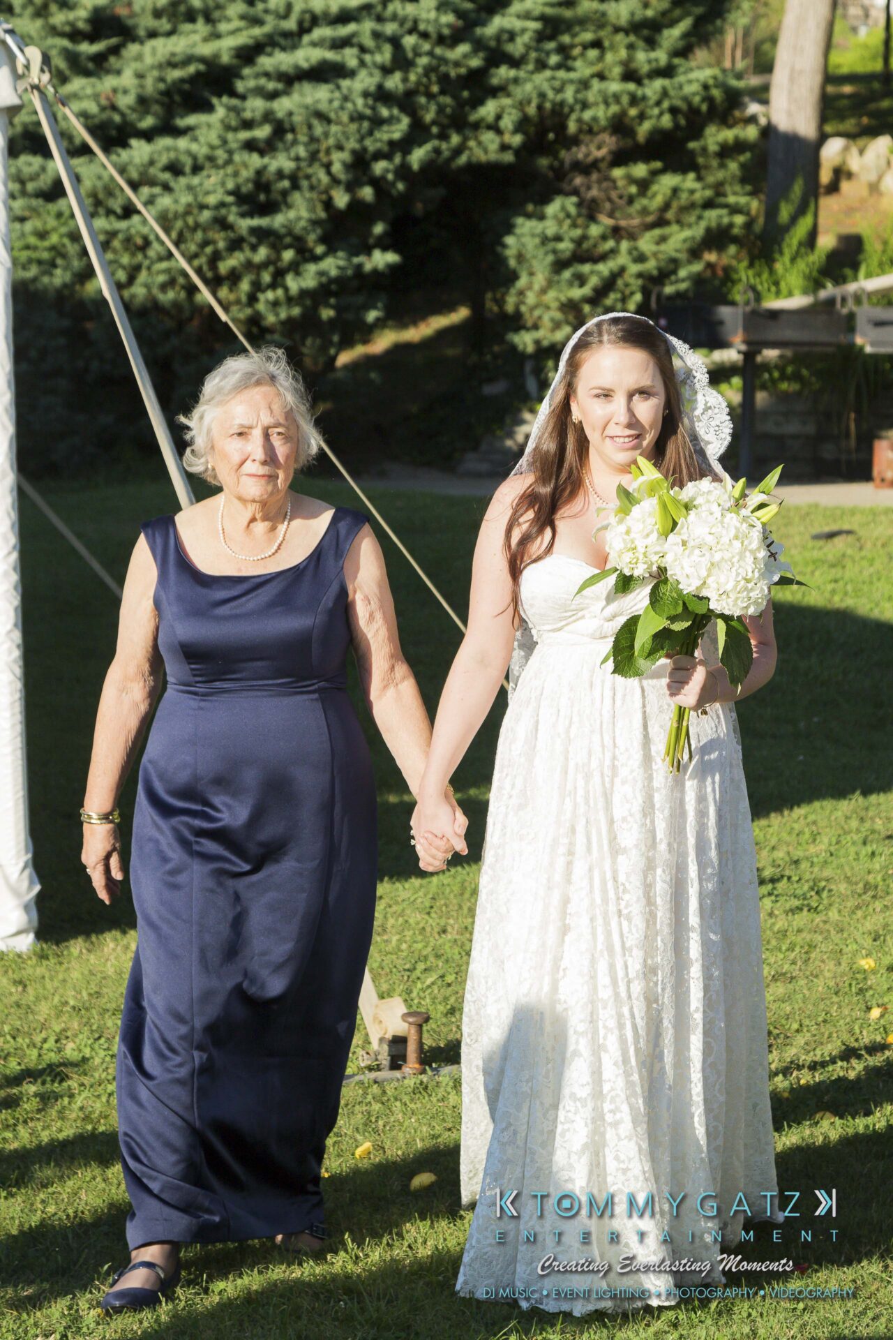 Ann-Marie-Alex-92317-CEREMONY-Low-Res-WM-35 - TOMMY GATZ ENTERTAINMENT A bride in a white dress walks hand in hand with an older woman in a navy dress outdoors on a sunny day.