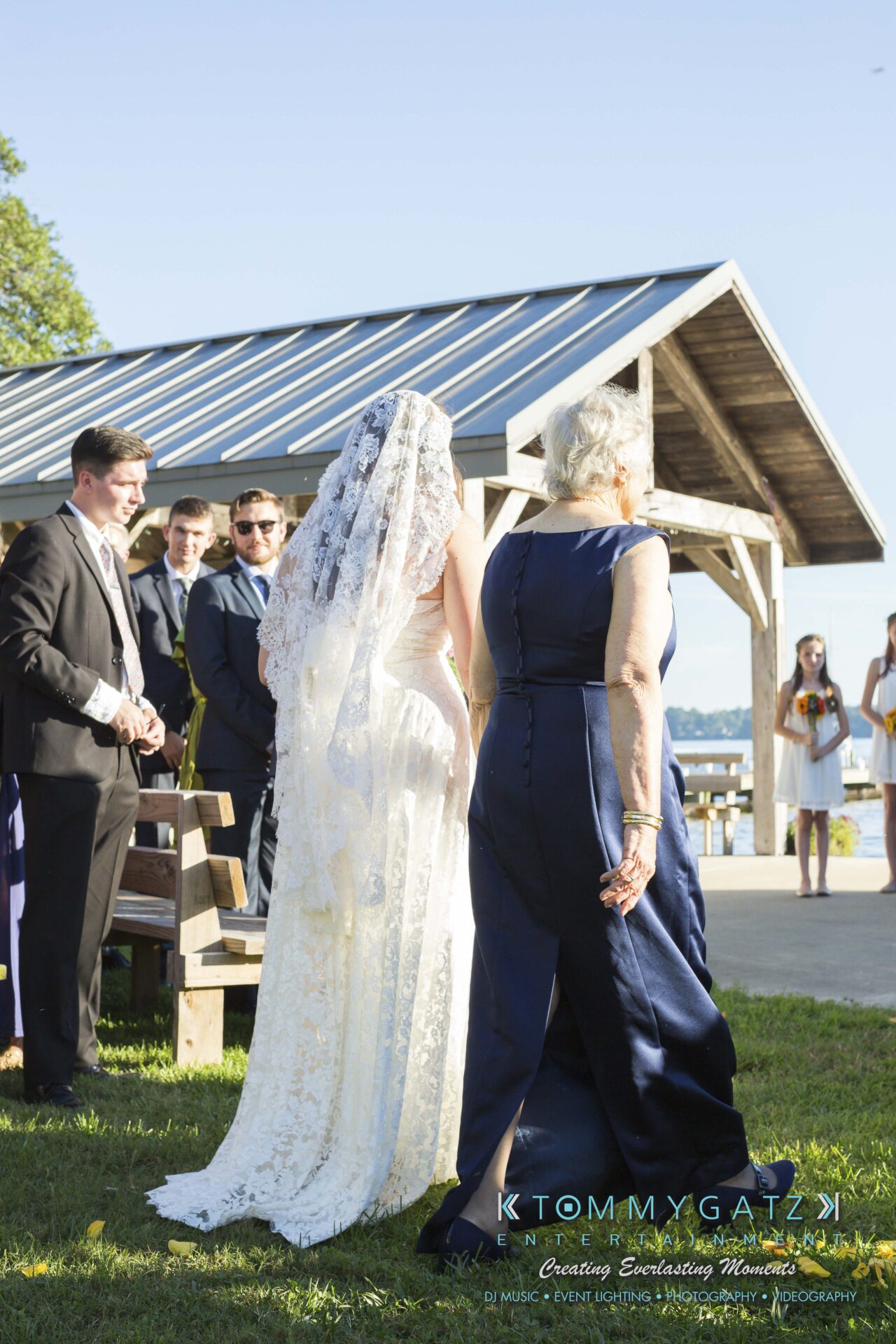 Ann-Marie-Alex-92317-CEREMONY-Low-Res-WM-37 - TOMMY GATZ ENTERTAINMENT A bride in a lace gown walks with an elderly woman towards a group of people in formal attire under a pavilion near a waterfront.