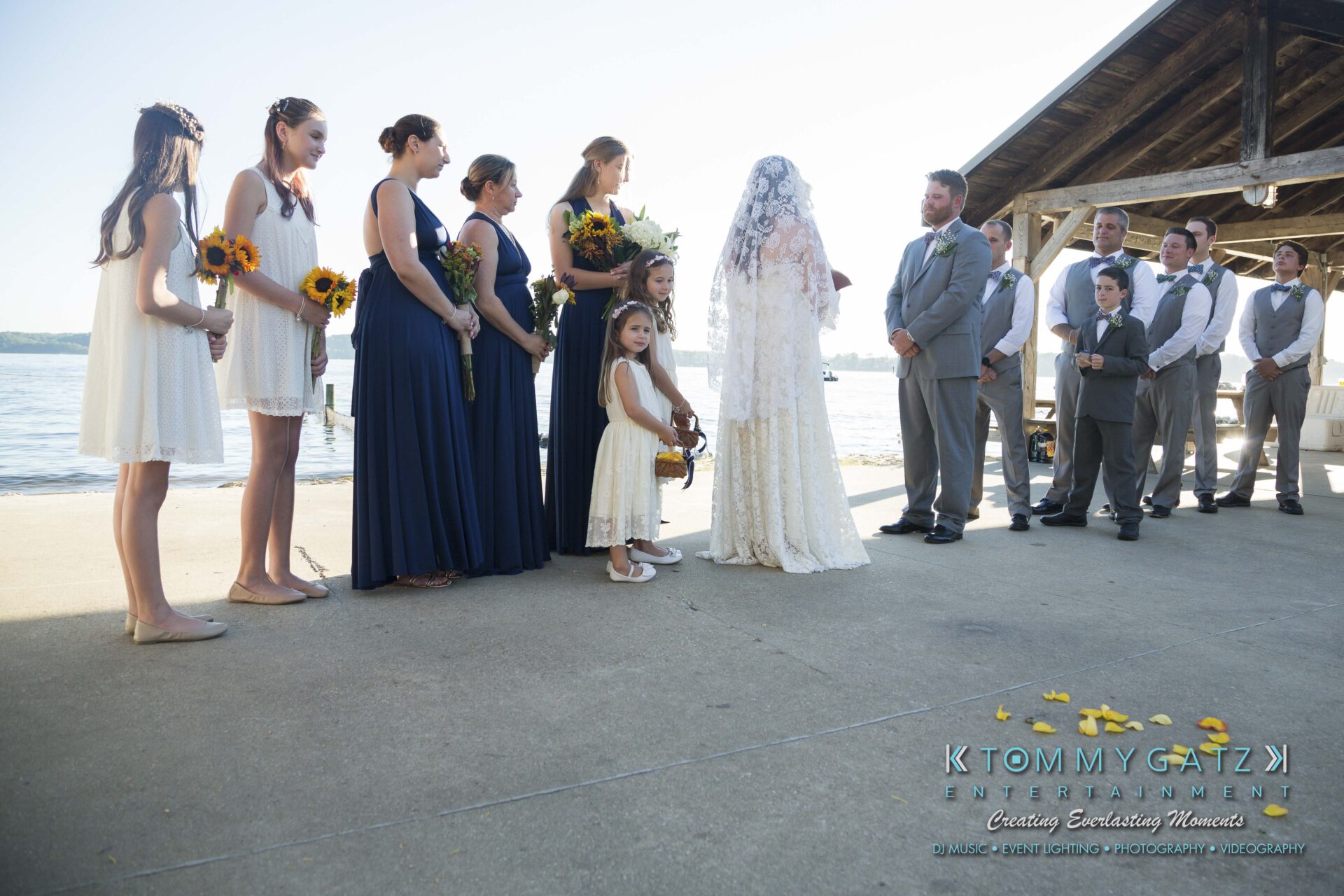 Ann-Marie-Alex-92317-CEREMONY-Low-Res-WM-44 - TOMMY GATZ ENTERTAINMENT A wedding party stands by a waterfront under a wooden pavilion. The bride and groom face each other, with bridesmaids and children holding sunflowers. Groomsmen are lined up beside them.