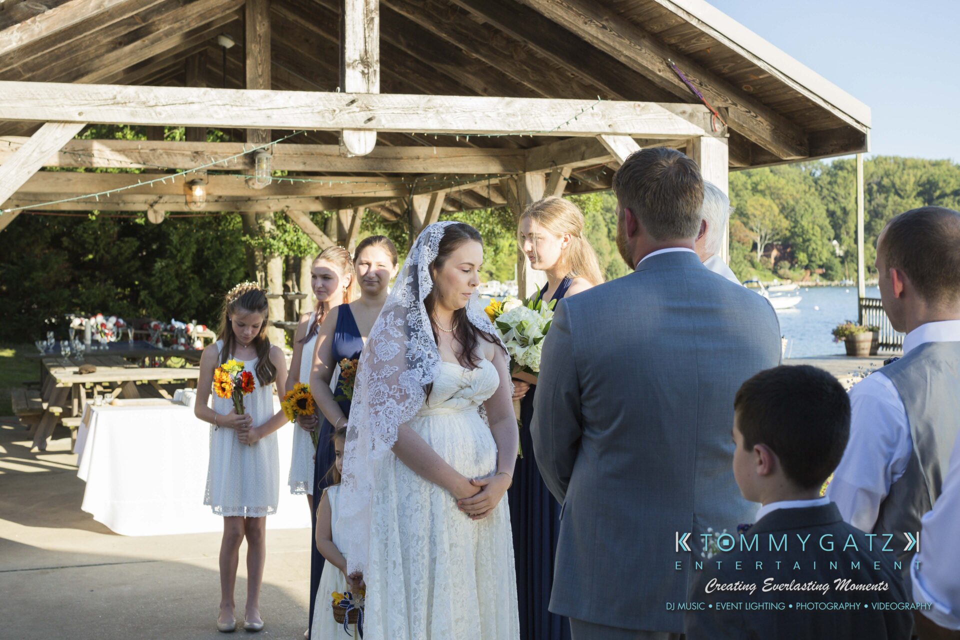 Ann-Marie-Alex-92317-CEREMONY-Low-Res-WM-45 - TOMMY GATZ ENTERTAINMENT A bride and groom stand at an outdoor wedding ceremony with bridesmaids and a flower girl by their side under a wooden gazebo.