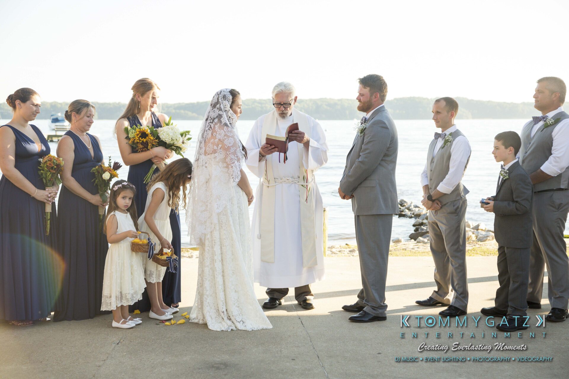 Ann-Marie-Alex-92317-CEREMONY-Low-Res-WM-51 - TOMMY GATZ ENTERTAINMENT A wedding ceremony by the water, with a bride and groom facing the officiant. Bridesmaids and groomsmen stand on either side, and two young flower girls are present.