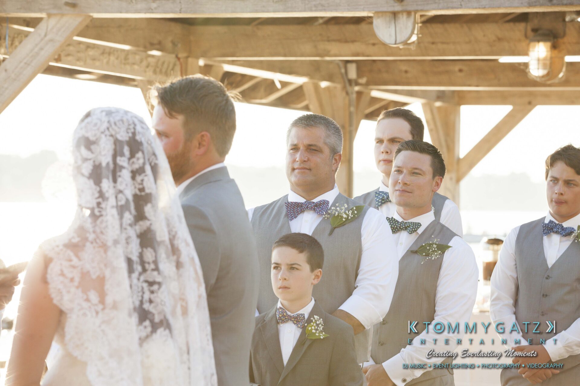 Ann-Marie-Alex-92317-CEREMONY-Low-Res-WM-58 - TOMMY GATZ ENTERTAINMENT A wedding ceremony with the bride in a lace veil and a group of groomsmen in gray vests and bow ties. The setting appears to be outdoors near a wooden structure.