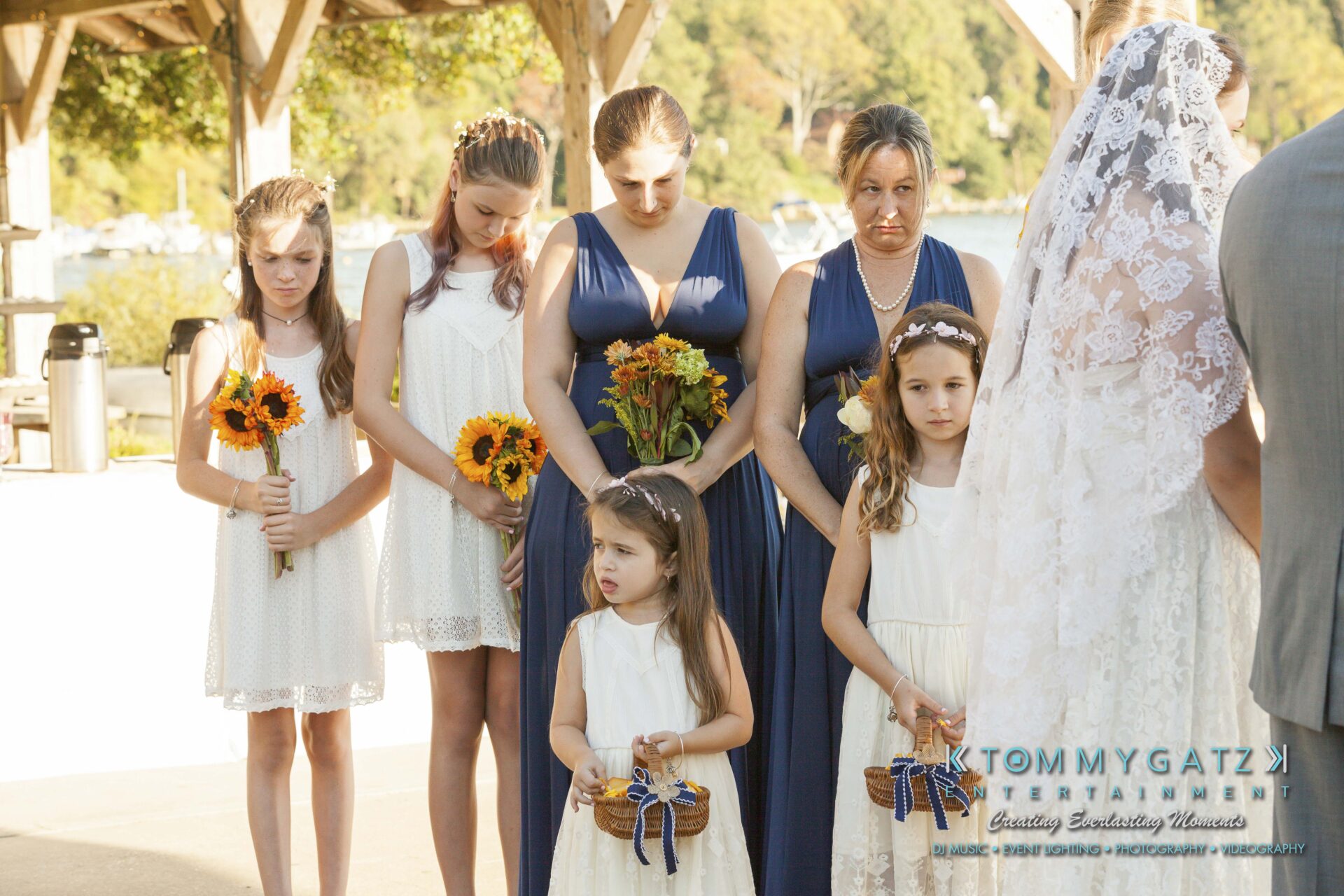 Ann-Marie-Alex-92317-CEREMONY-Low-Res-WM-66 - TOMMY GATZ ENTERTAINMENT Wedding party lined up outdoors: women in blue dresses, girls in white holding flowers and baskets. Bride in veil partly visible on the right.
