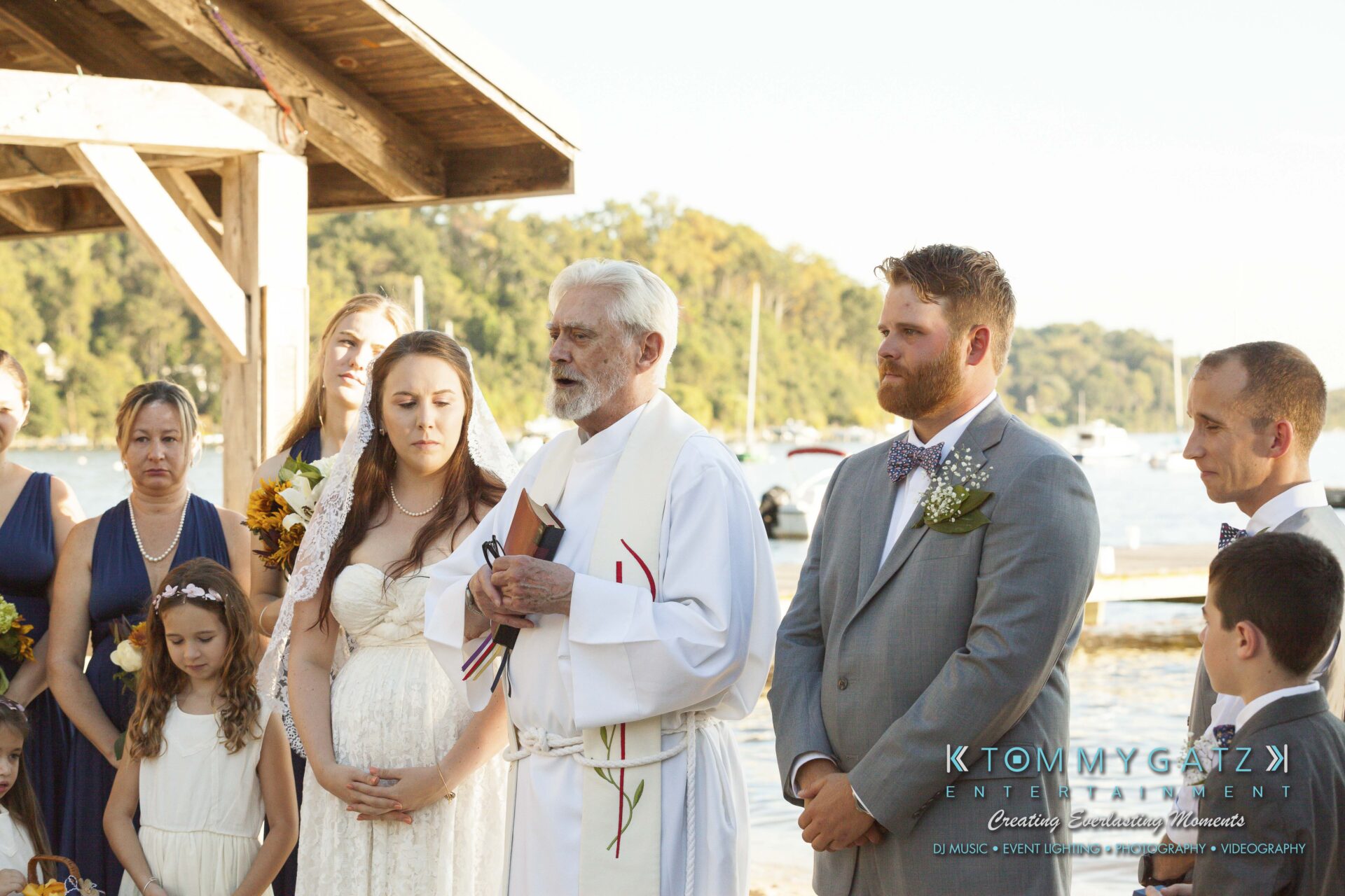Ann-Marie-Alex-92317-CEREMONY-Low-Res-WM-70 - TOMMY GATZ ENTERTAINMENT A wedding ceremony by a lakeside, featuring a bride and groom with a priest. Bridesmaids and guests stand nearby.