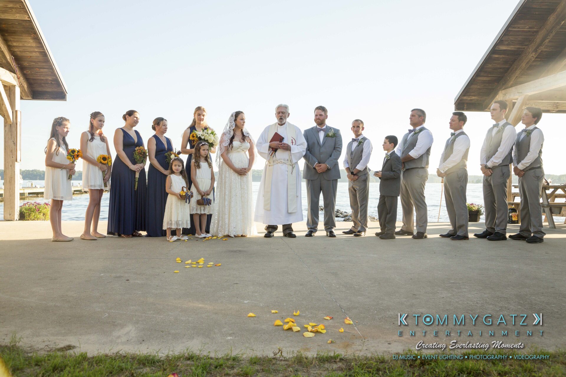 Ann-Marie-Alex-92317-CEREMONY-Low-Res-WM-72 - TOMMY GATZ ENTERTAINMENT A wedding ceremony by the water with a bride, groom, officiant, and wedding party standing under a wooden structure.