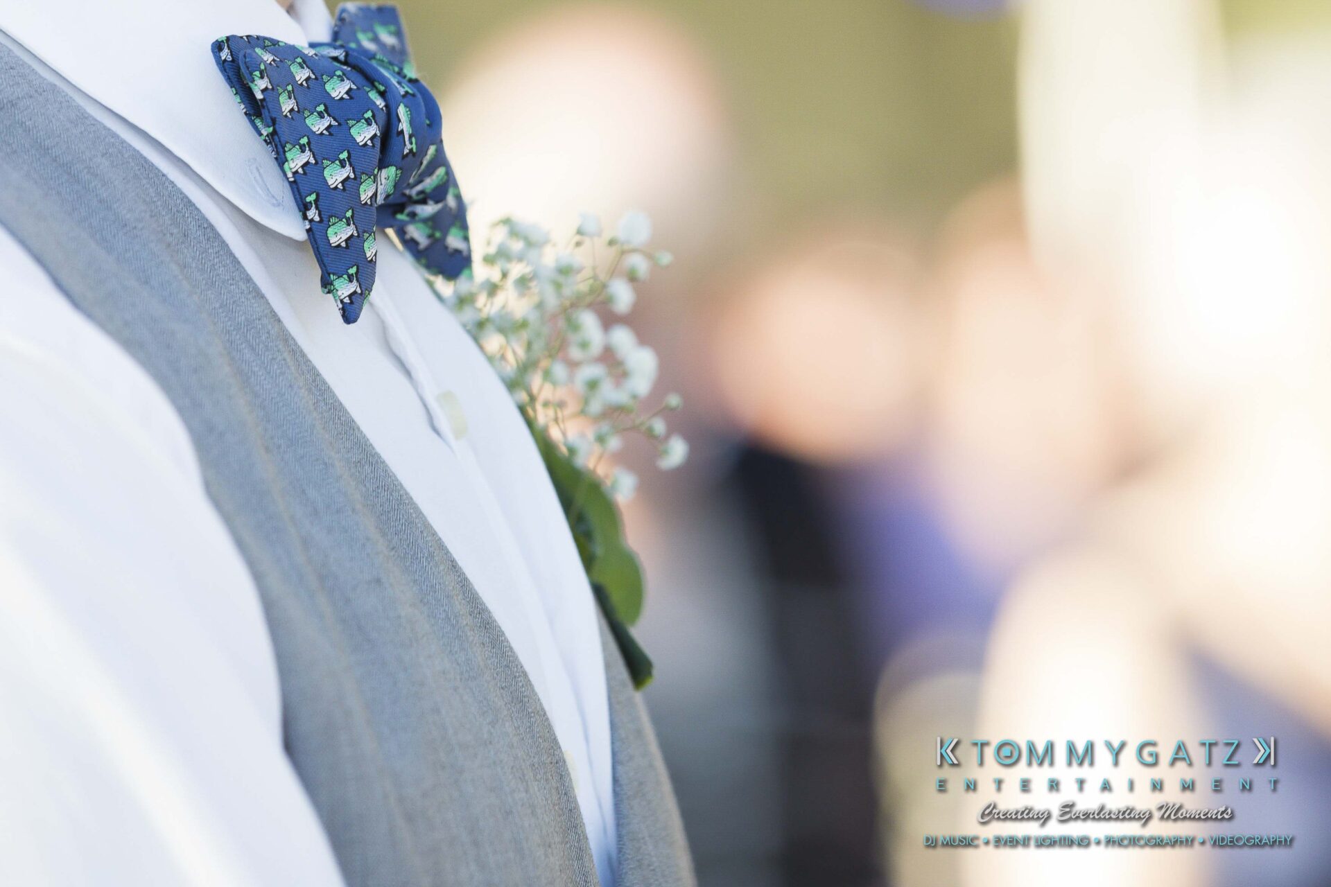 Ann-Marie-Alex-92317-CEREMONY-Low-Res-WM-87 - TOMMY GATZ ENTERTAINMENT Close-up of a person wearing a blue bow tie with a floral pattern, a white shirt, and a gray vest. A sprig of flowers is in the vest pocket.