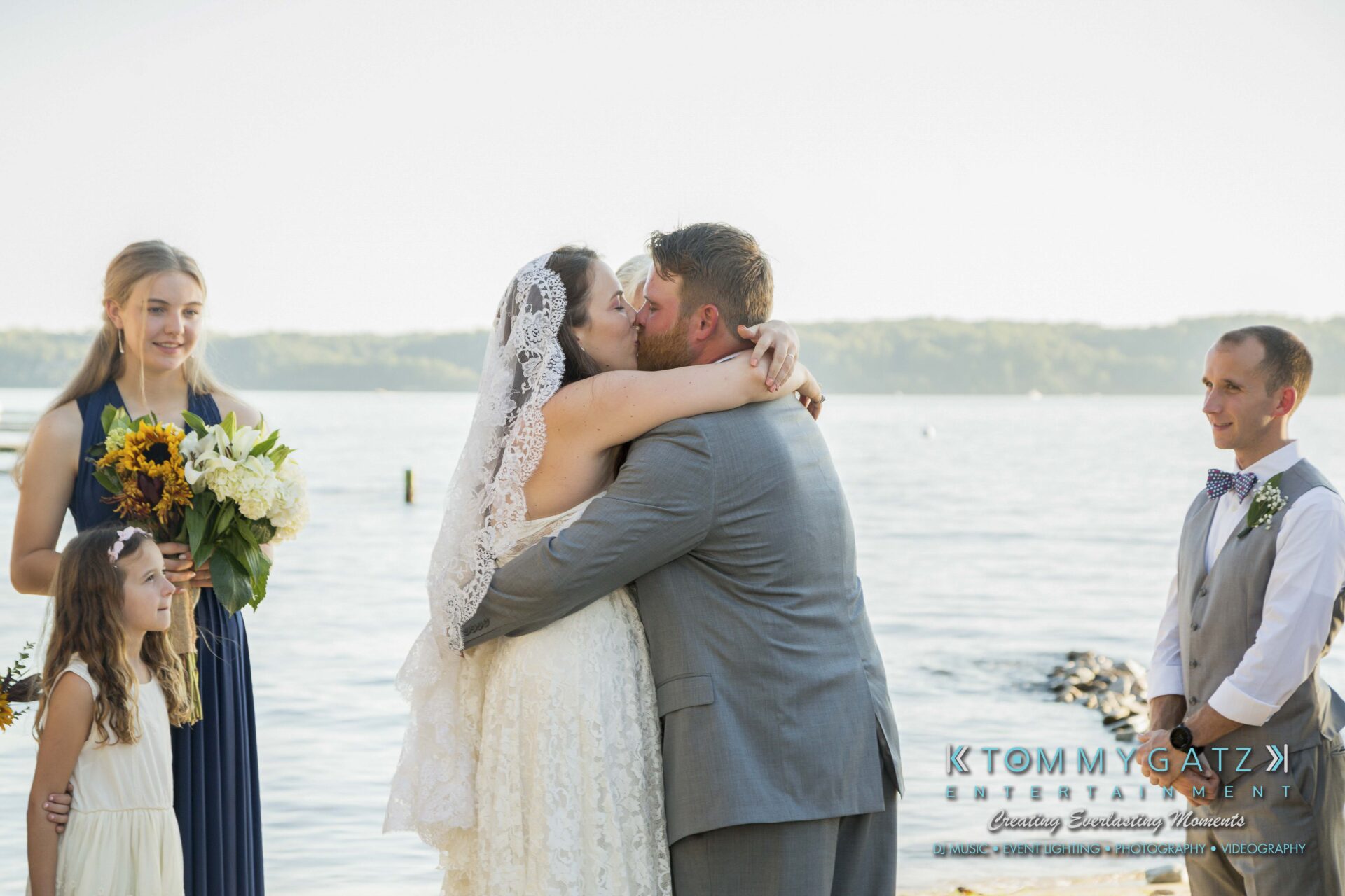 Ann-Marie-Alex-92317-CEREMONY-Low-Res-WM-94 - TOMMY GATZ ENTERTAINMENT A couple kisses during a lakeside wedding ceremony. Bridesmaids and the officiant stand nearby.