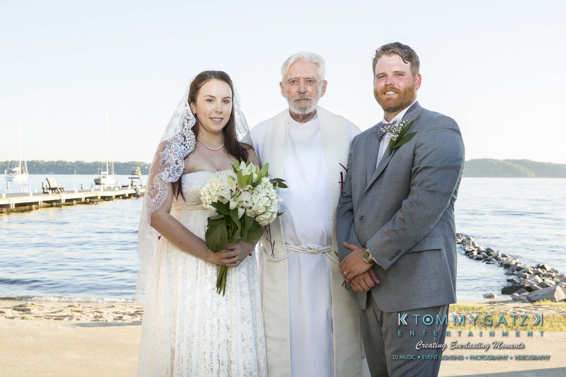 Ann-Marie-Alex-92317-FORMALS-Low-Res-WM-1 - TOMMY GATZ ENTERTAINMENT Bride and groom pose with officiant by a lakeside, under clear skies.