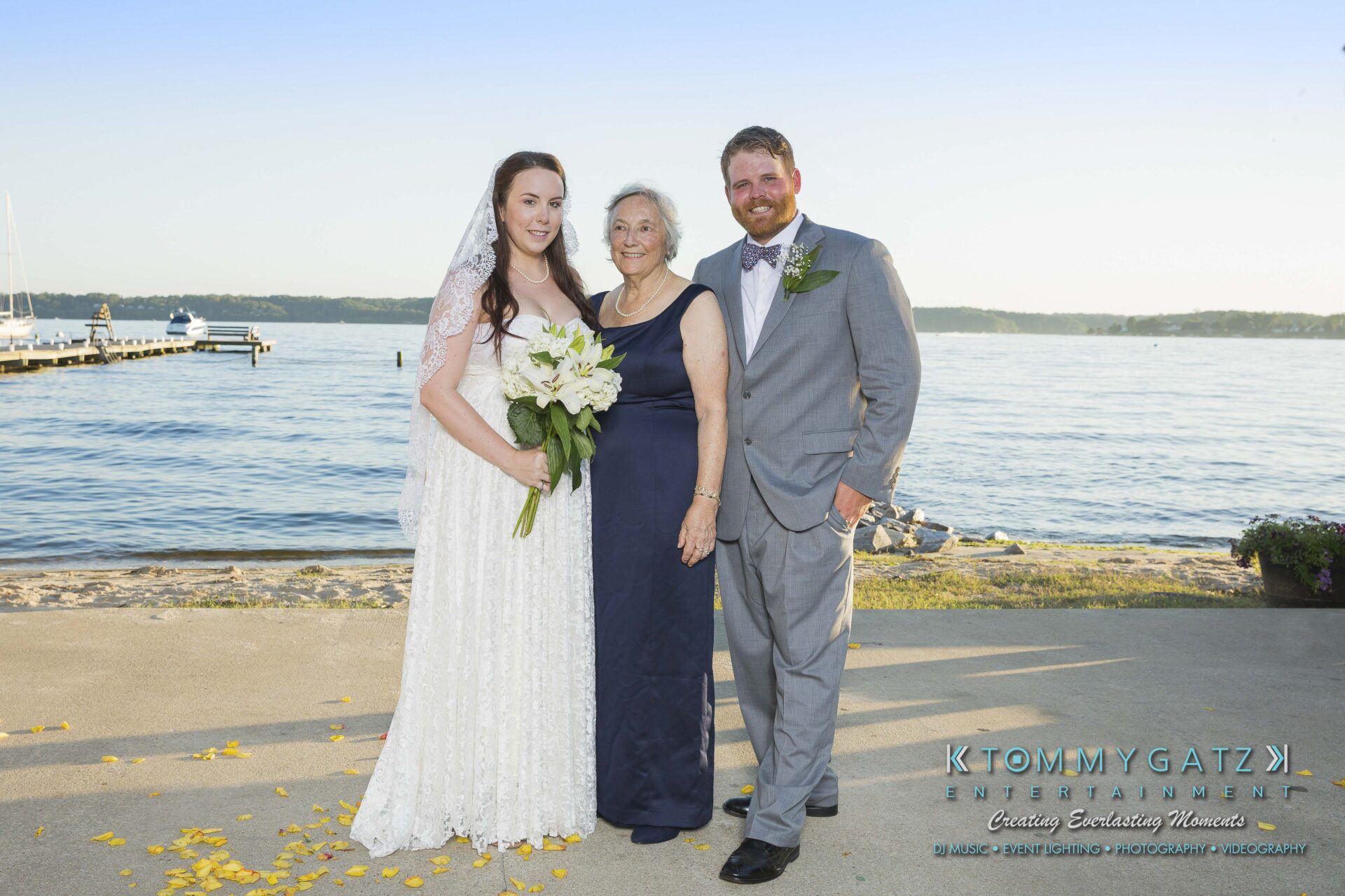 Ann-Marie-Alex-92317-FORMALS-Low-Res-WM-10 - TOMMY GATZ ENTERTAINMENT A bride, an elderly woman, and a groom stand smiling by a waterfront. The bride holds a bouquet, and the groom is in a gray suit.