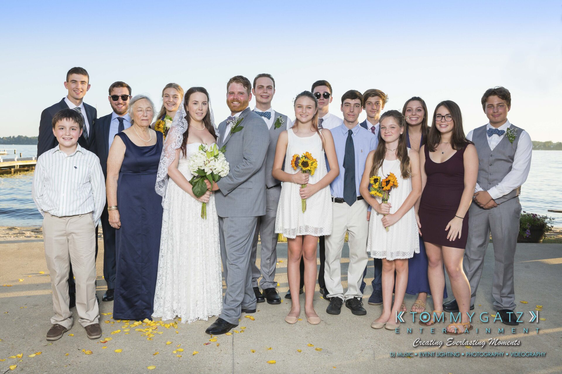 Ann-Marie-Alex-92317-FORMALS-Low-Res-WM-14 - TOMMY GATZ ENTERTAINMENT A group photo of wedding guests, including the bride and groom, standing outdoors by a body of water. Some hold bouquets of sunflowers and white flowers.