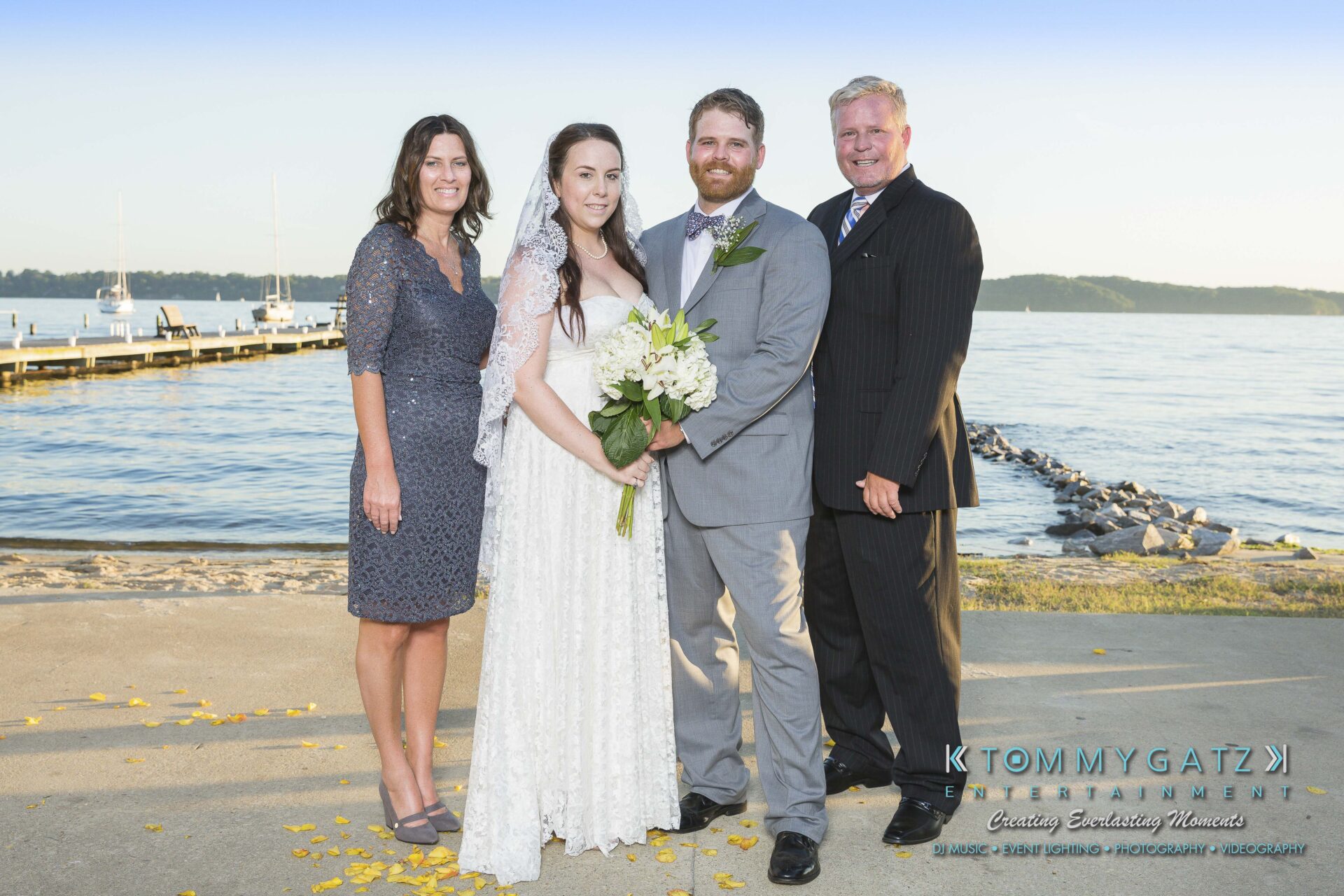 Ann-Marie-Alex-92317-FORMALS-Low-Res-WM-17 - TOMMY GATZ ENTERTAINMENT A wedding couple stands with two others by a waterfront, smiling at the camera. The bride holds a bouquet. There is a pier and calm water in the background.