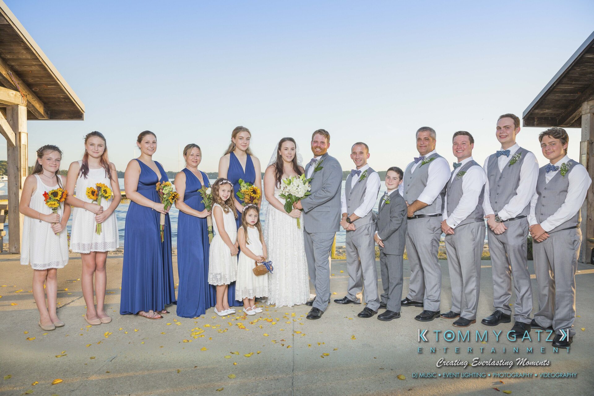Ann-Marie-Alex-92317-FORMALS-Low-Res-WM-20 - TOMMY GATZ ENTERTAINMENT A wedding party poses outdoors. The group includes the bride, groom, bridesmaids in blue dresses, and groomsmen in gray suits. The setting is sunny with a clear blue sky.