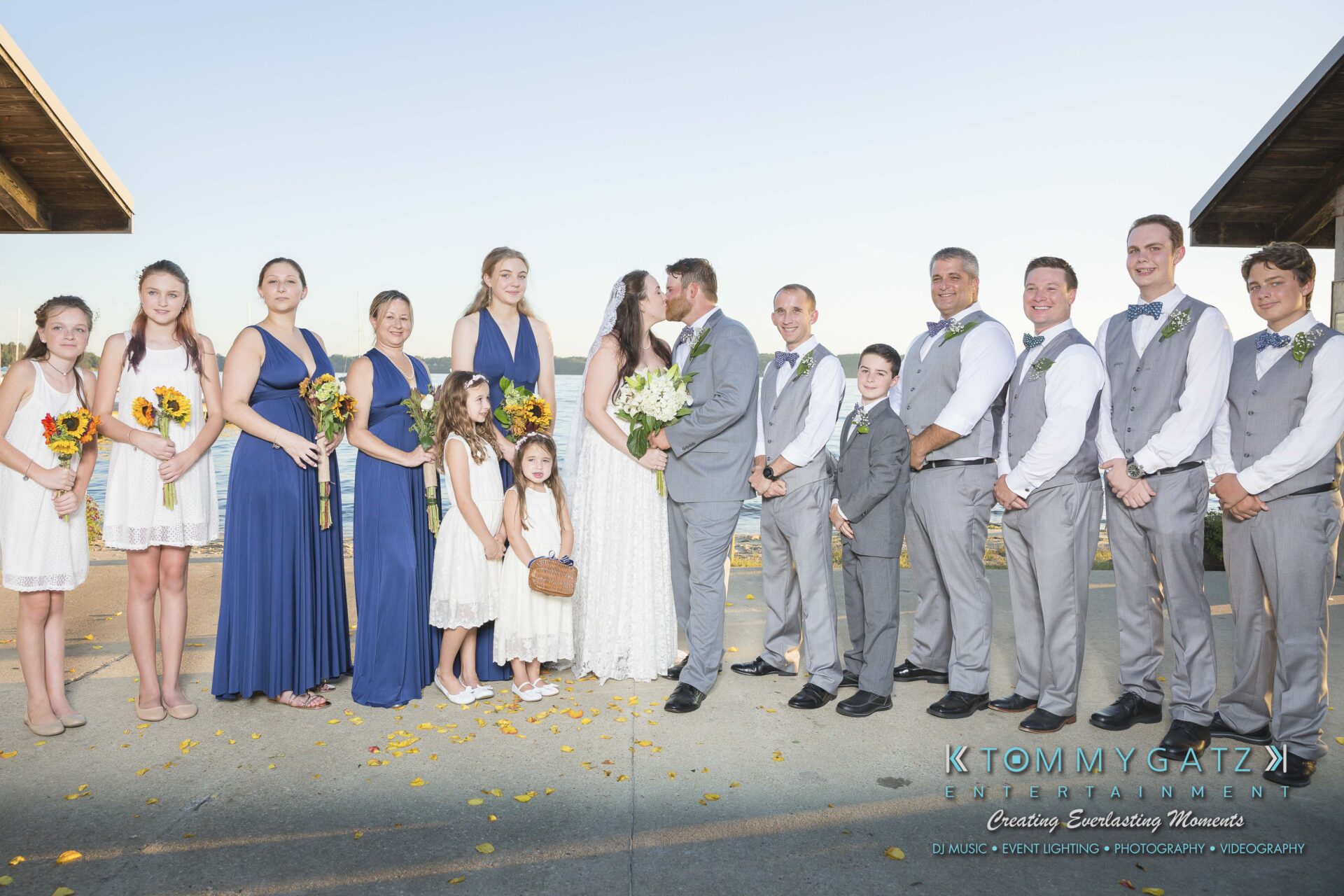 Ann-Marie-Alex-92317-FORMALS-Low-Res-WM-21 - TOMMY GATZ ENTERTAINMENT Wedding party stands in a line outdoors, with a couple kissing in the center, surrounded by bridesmaids in blue and white dresses and groomsmen in gray suits.
