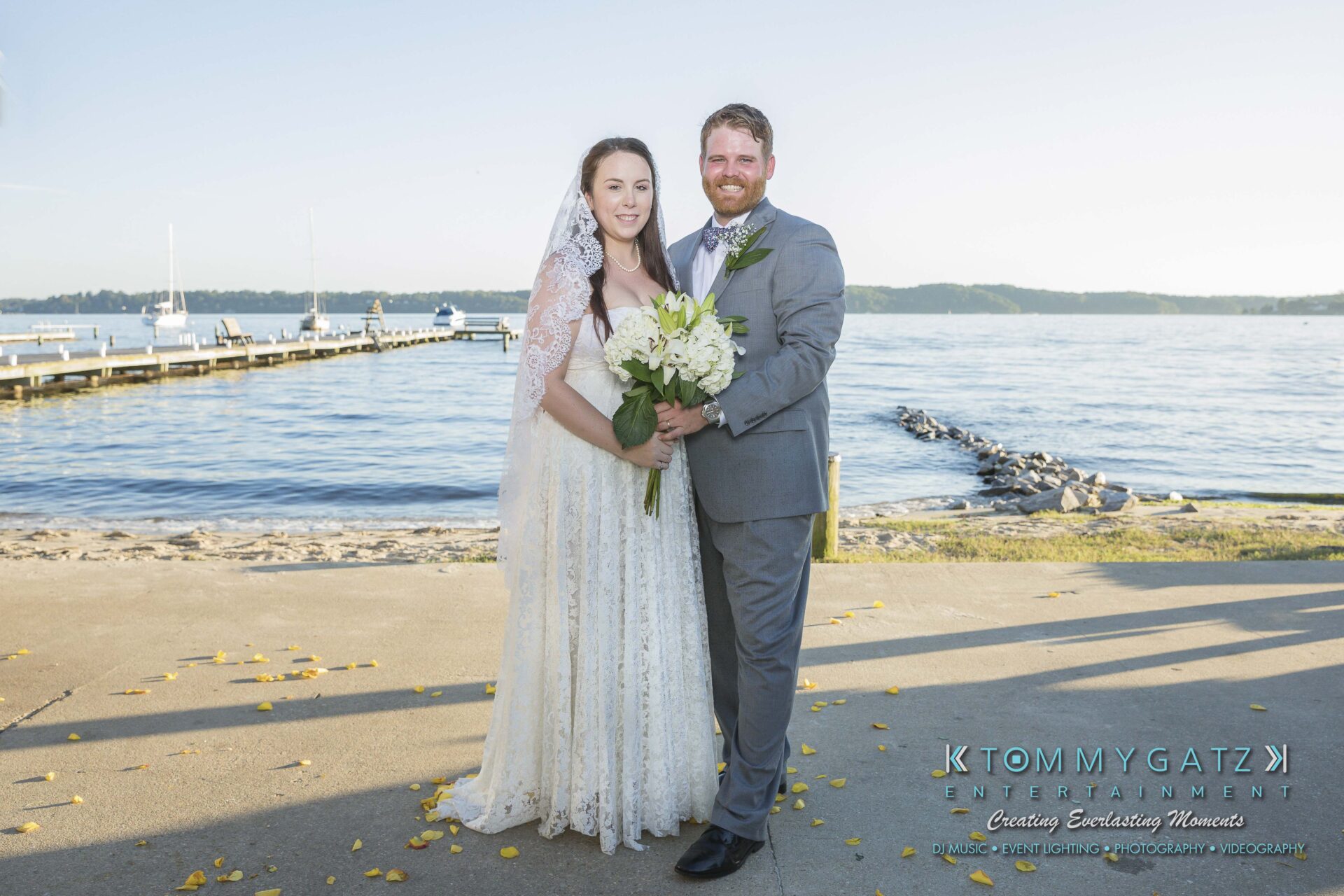 Ann-Marie-Alex-92317-FORMALS-Low-Res-WM-24 - TOMMY GATZ ENTERTAINMENT A bride and groom stand together by the waterfront on a sunny day, the bride holding a bouquet of white flowers.
