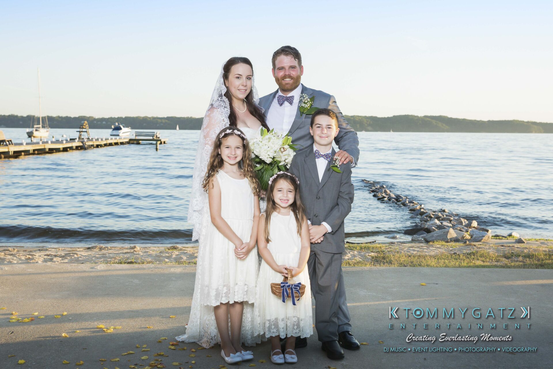Ann-Marie-Alex-92317-FORMALS-Low-Res-WM-25 - TOMMY GATZ ENTERTAINMENT A family poses by a lakeside, with a bride and groom standing behind three children. A pier extends into the water, and distant hills are visible in the background.