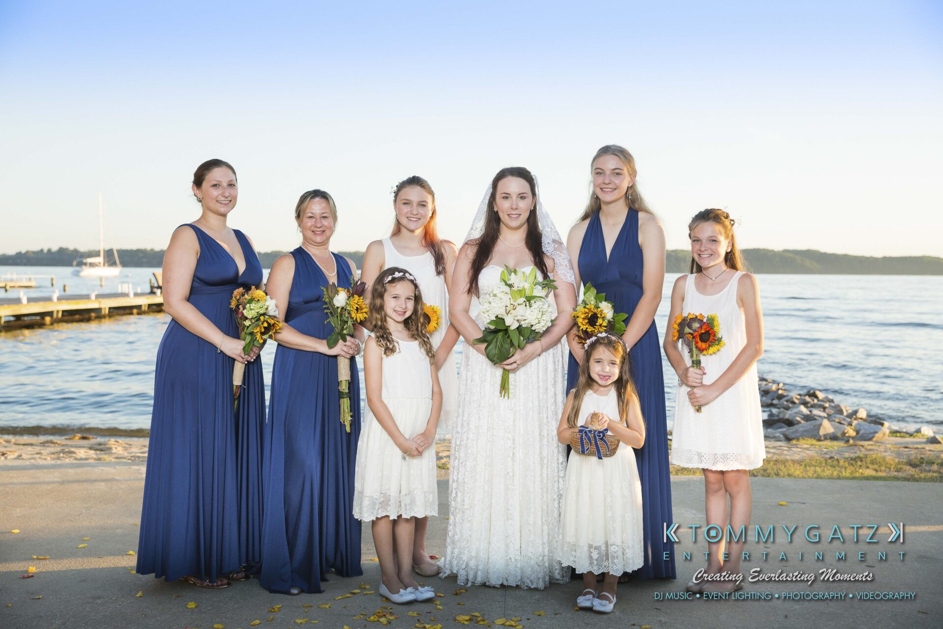 Ann-Marie-Alex-92317-FORMALS-Low-Res-WM-27 - TOMMY GATZ ENTERTAINMENT A bride stands with six bridesmaids and two flower girls in white and blue dresses by a lakeside.