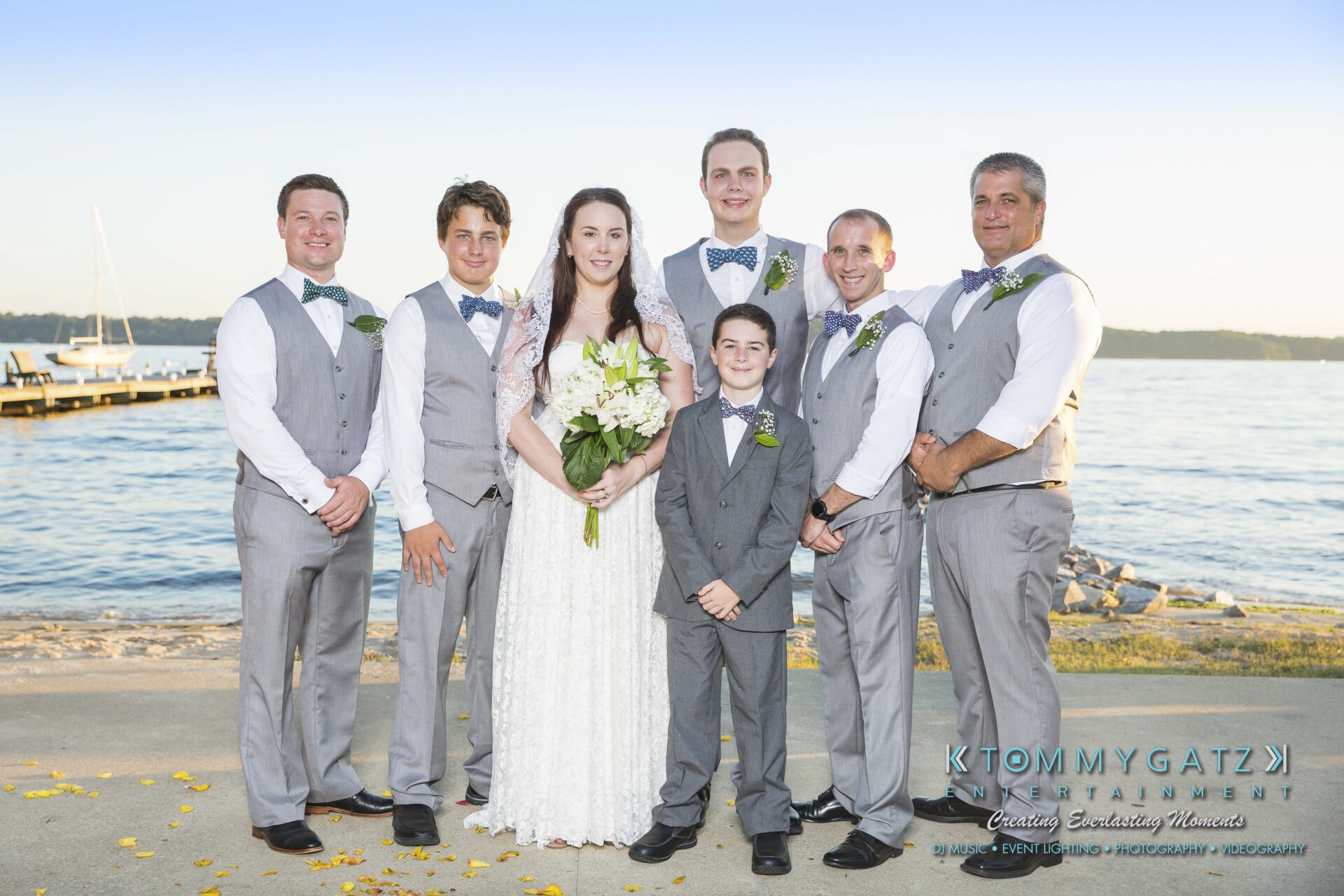 Ann-Marie-Alex-92317-FORMALS-Low-Res-WM-28 - TOMMY GATZ ENTERTAINMENT A wedding party poses by the water, featuring a bride in a white dress holding flowers, a young boy, and five men in gray vests and bow ties. The background includes a dock and calm water.